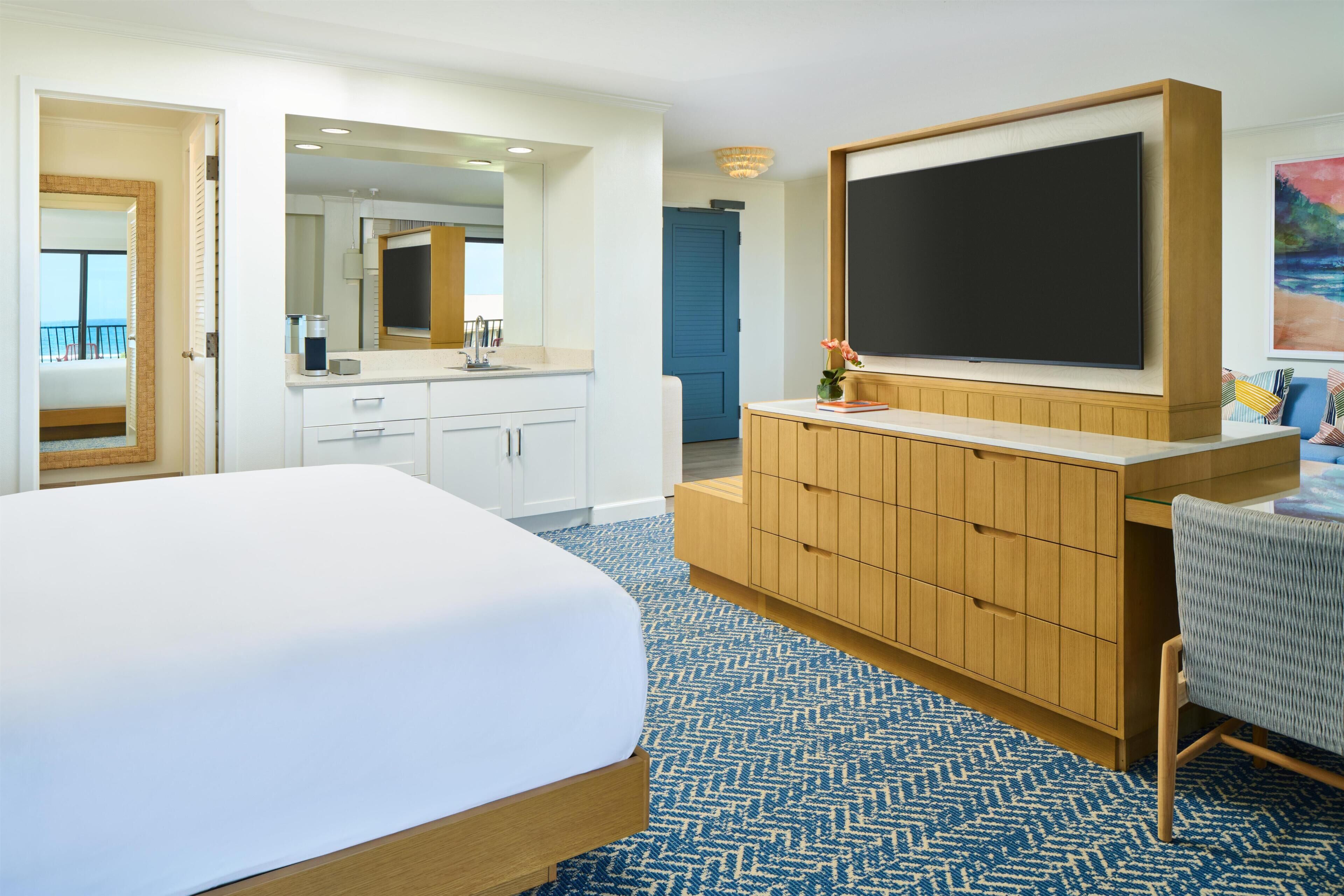 A hotel room with a large bed, a wooden dresser, a flat-screen TV, a bathroom with a mirror, and a view of the outside.