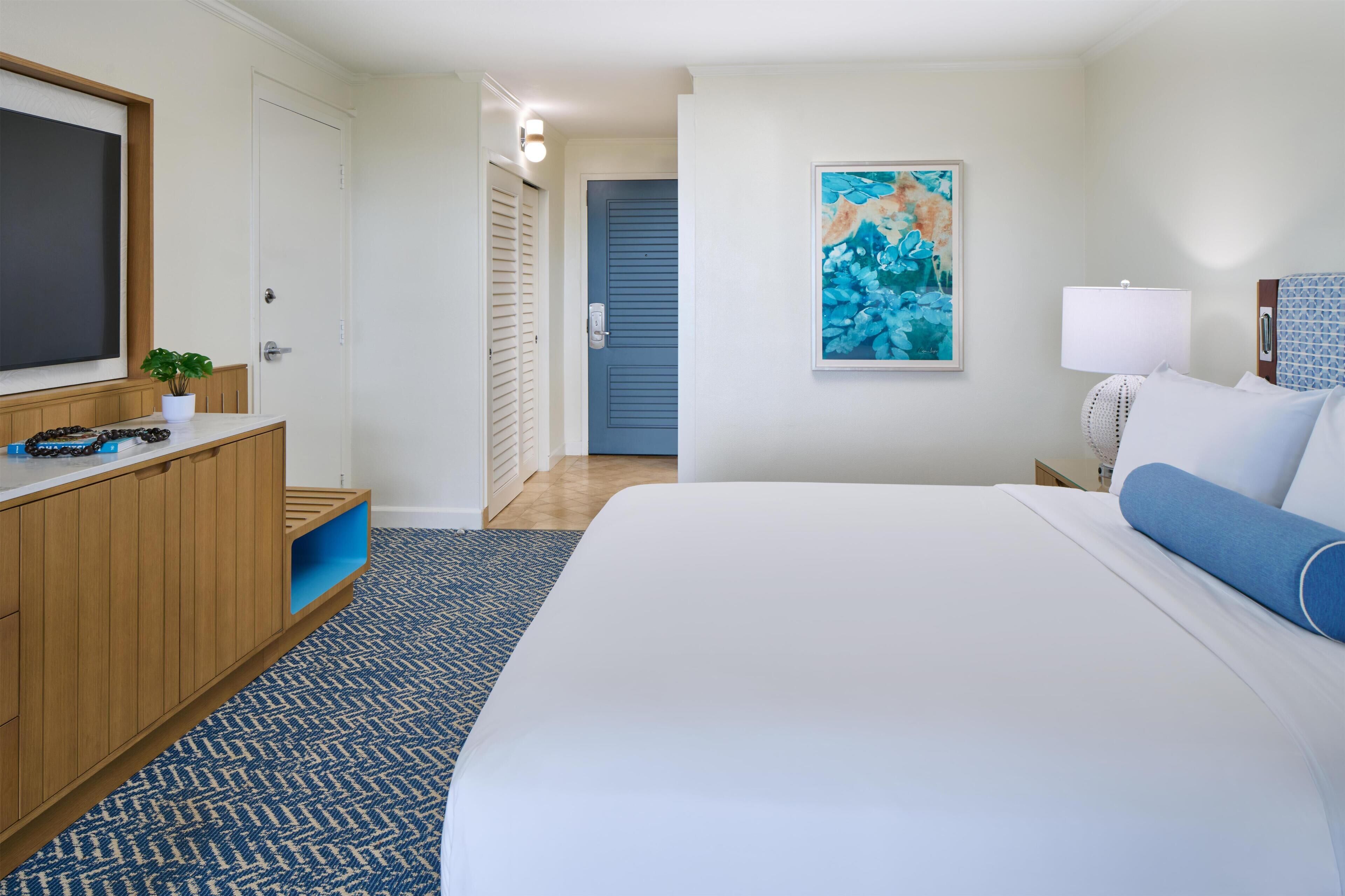 A hotel room with a bed, a TV, a painting, a lamp, and a wooden dresser.