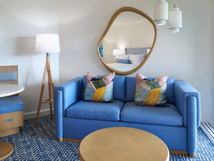 A modern hotel room with a blue sofa, a round wooden table, a mirror, and a patterned floor.