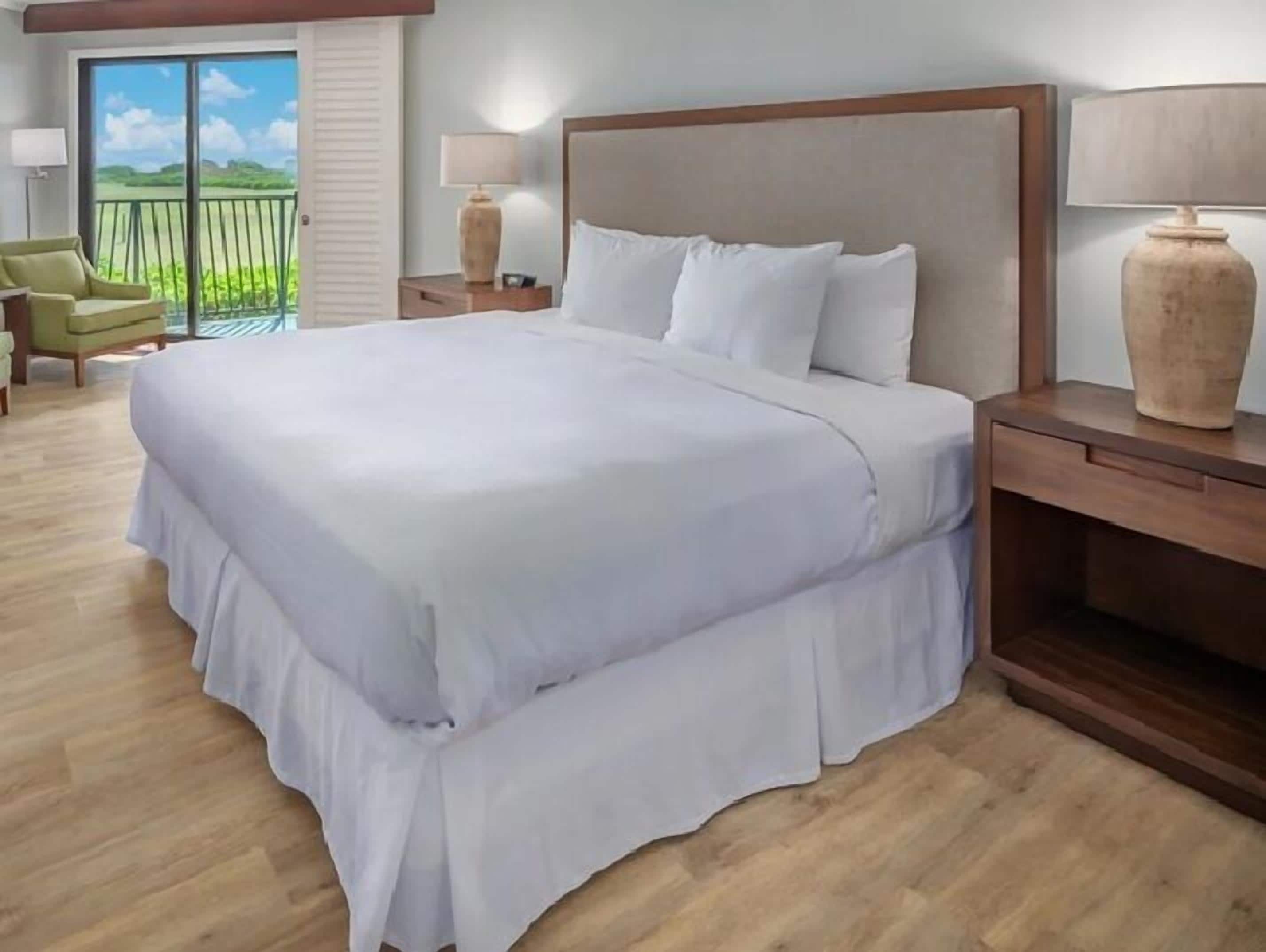 A hotel room with a large bed, a nightstand, a lamp, a chair, and a view of the outdoors.