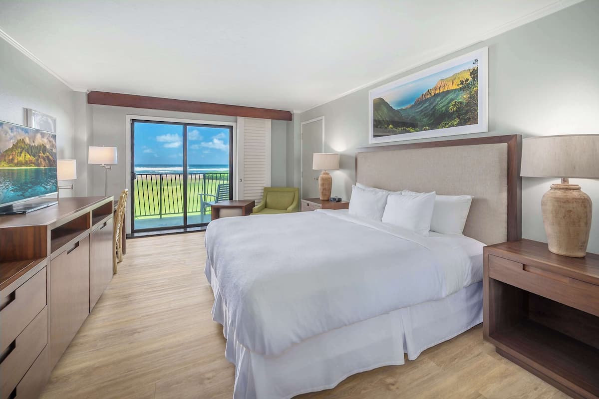 A hotel room with a large bed, a desk, a chair, a nightstand, and a view of the ocean through a sliding door.