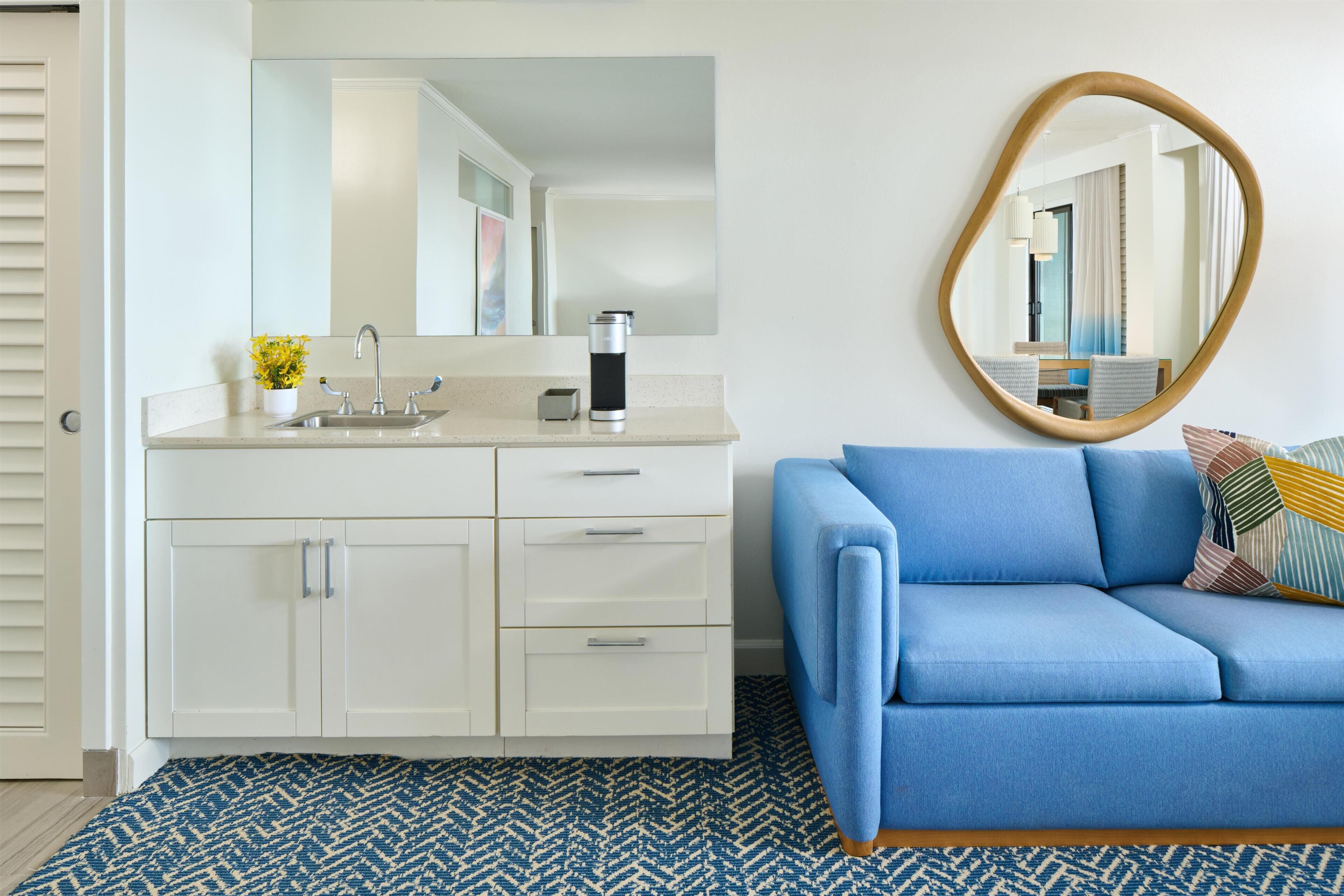Un baño moderno con tocador blanco, lavamanos, espejo y cafetera. En la zona de estar contigua, hay un sofá azul con cojines estampados.