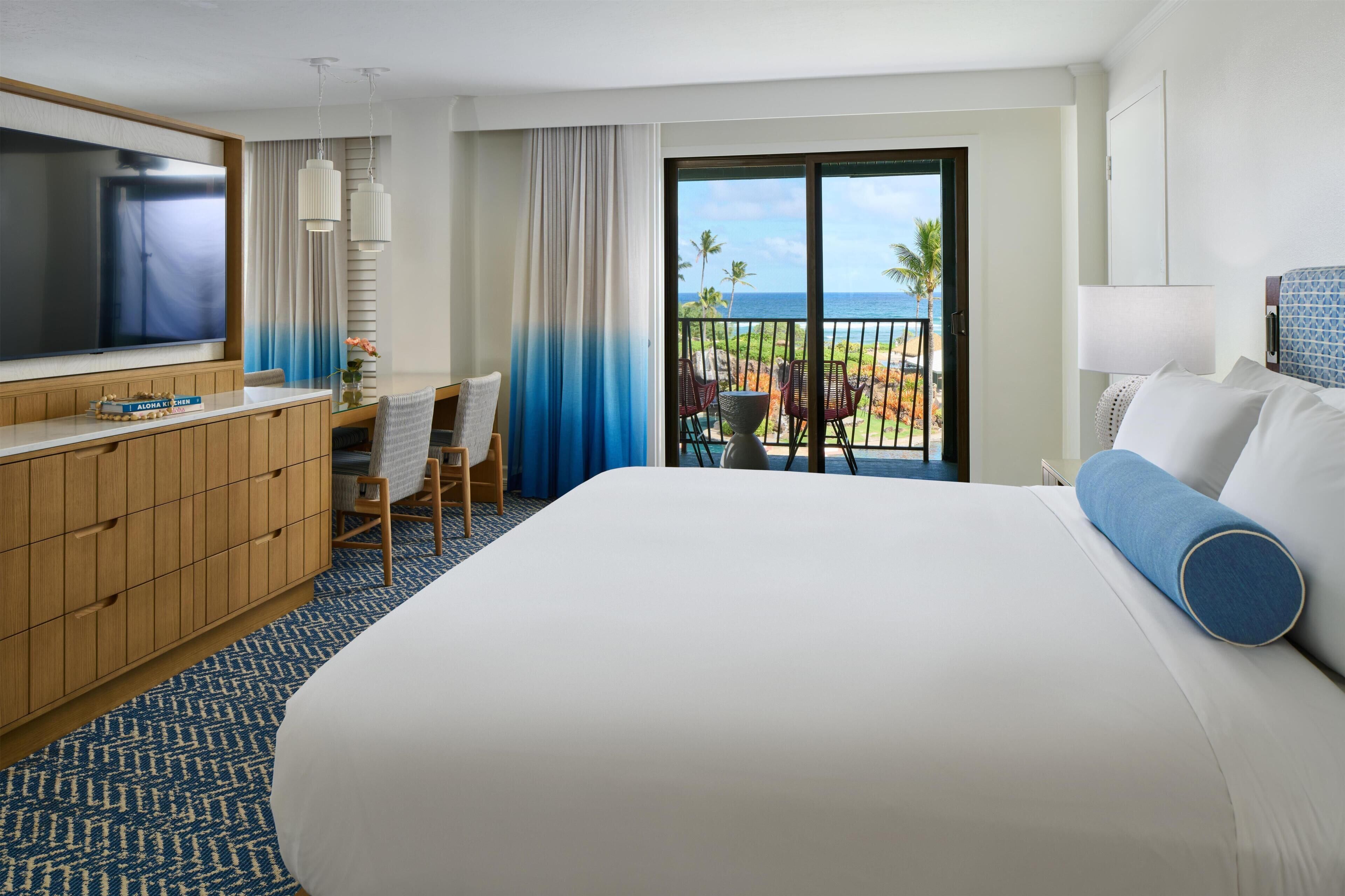 A hotel room with a large bed, a desk with a chair, a TV, and a balcony with a view of the ocean.