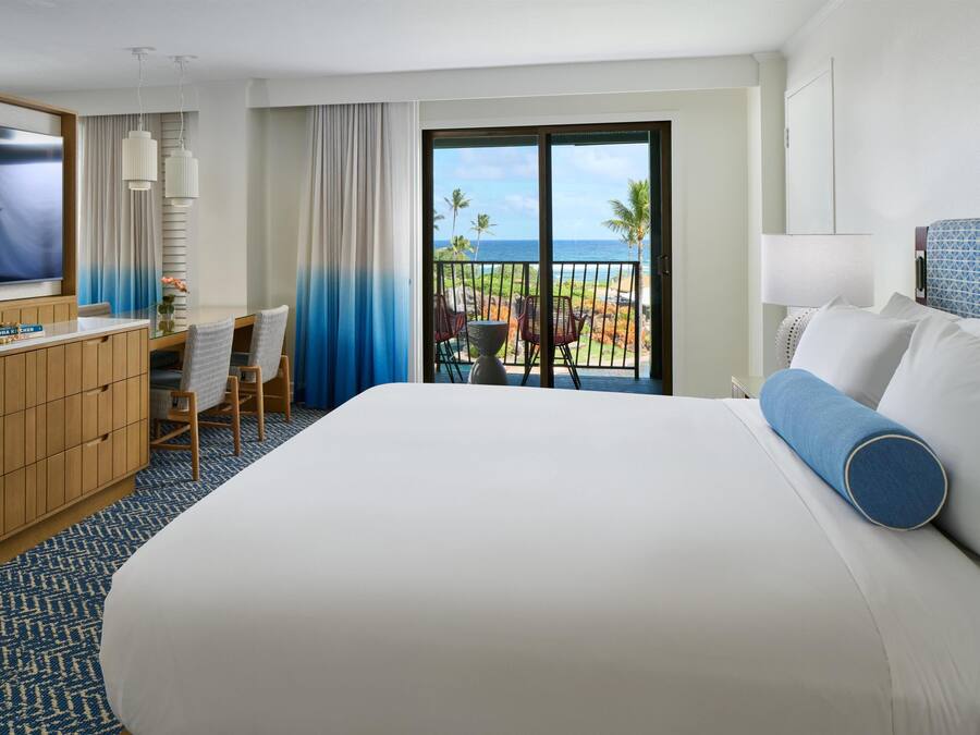 A hotel room with a large bed, a desk with a chair, a TV, and a balcony with a view of the ocean.