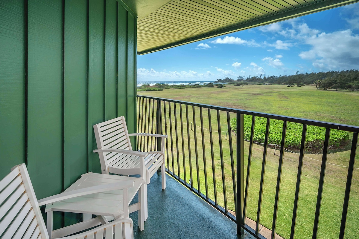 A balcony with two white chairs and a table, overlooking a grassy field and a distant shoreline.