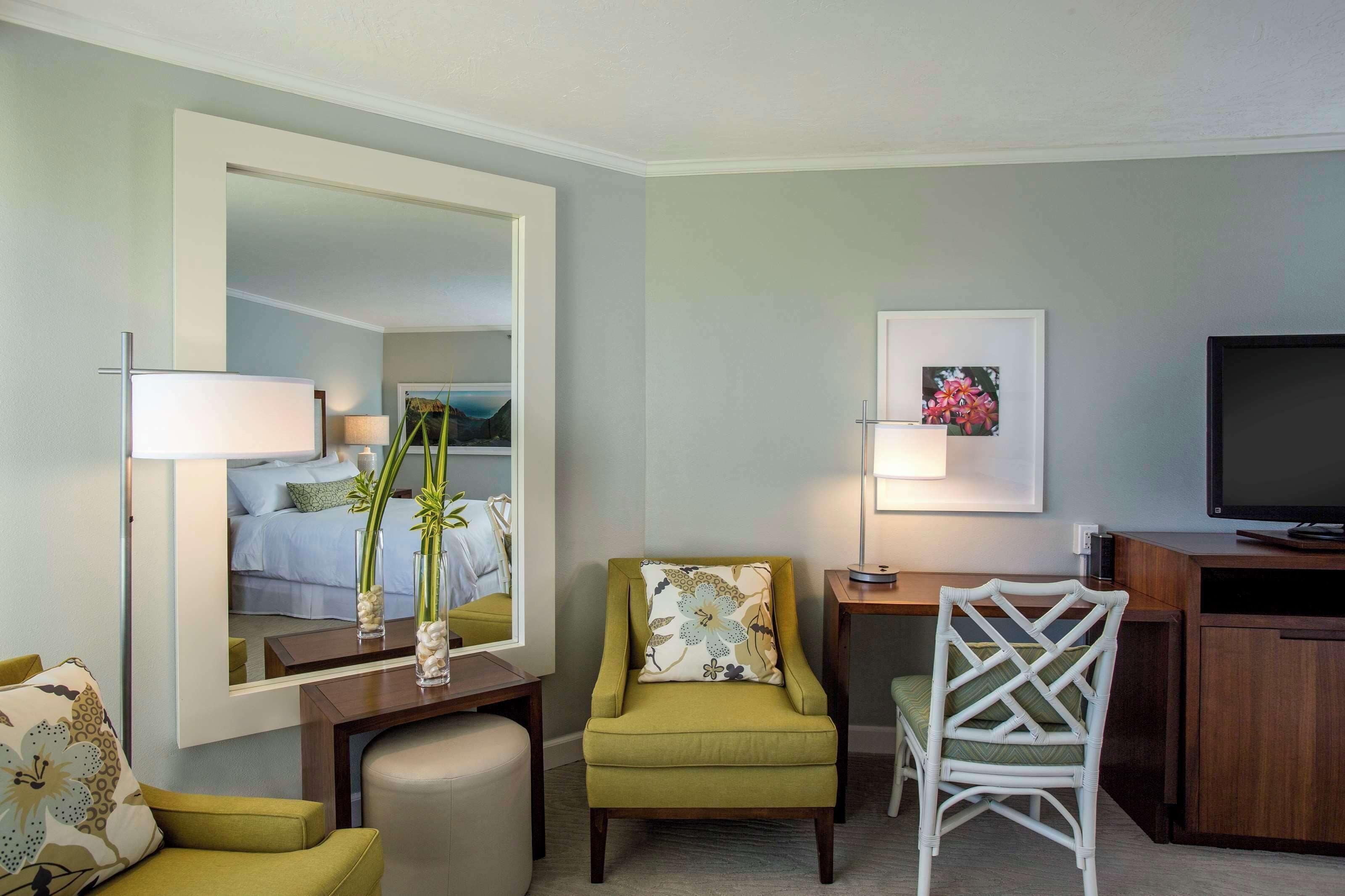 Habitación de hotel con cama, espejo, escritorio con lámpara, televisión y dos sillones.