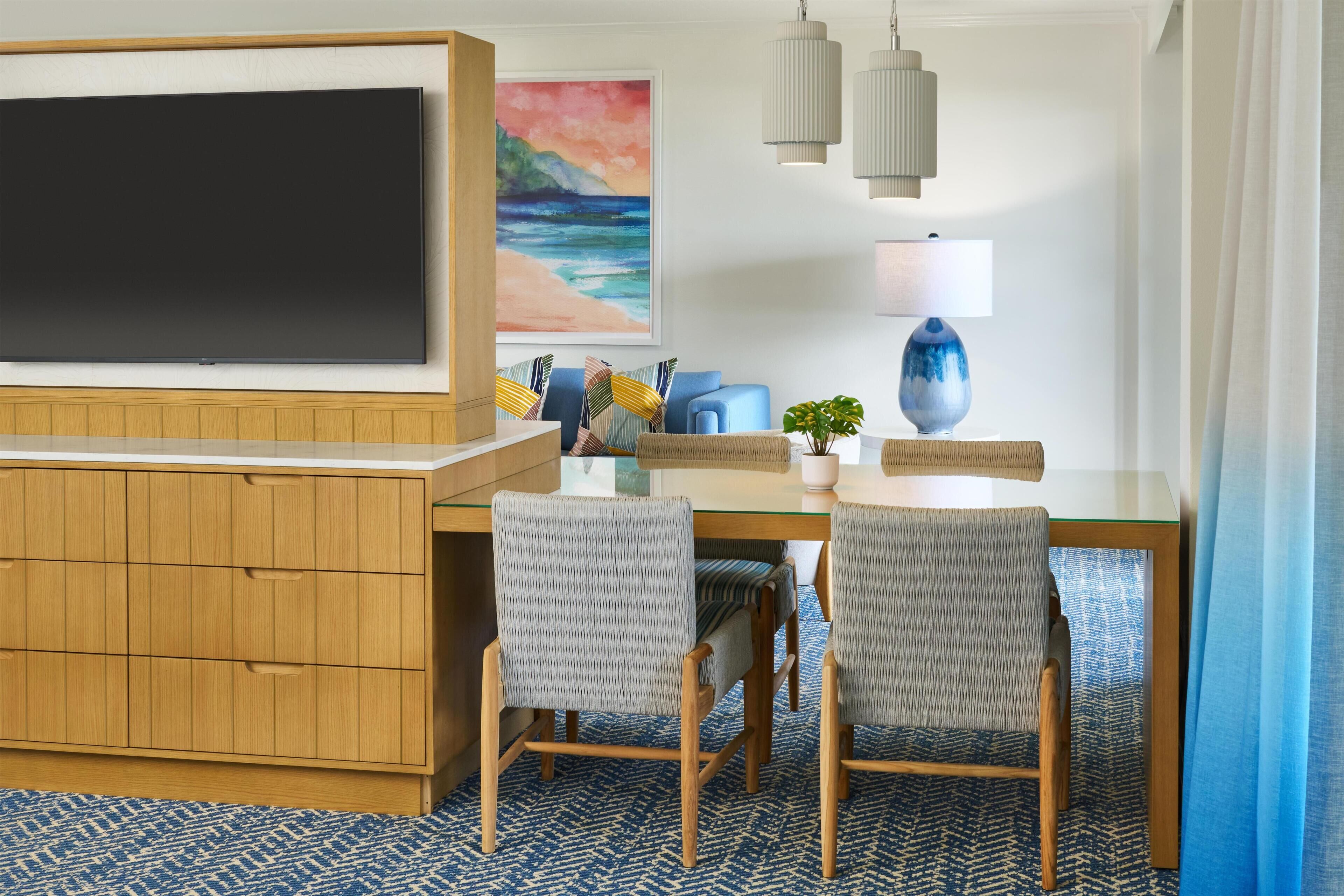 A hotel room with a flat-screen TV, a painting of a beach, a dining table with chairs, a lamp, and a plant.