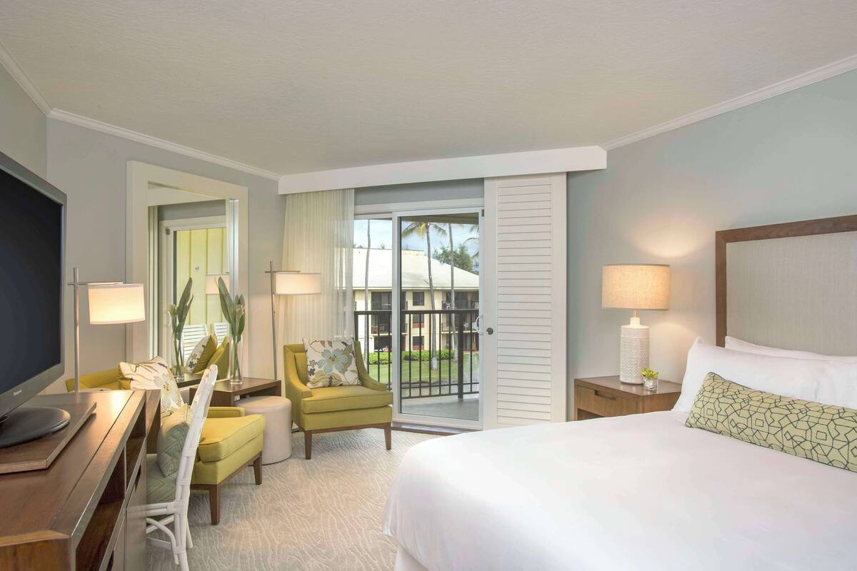 A hotel room with a bed, a desk with a TV, a chair, a nightstand with a lamp, and a view of the outdoors.