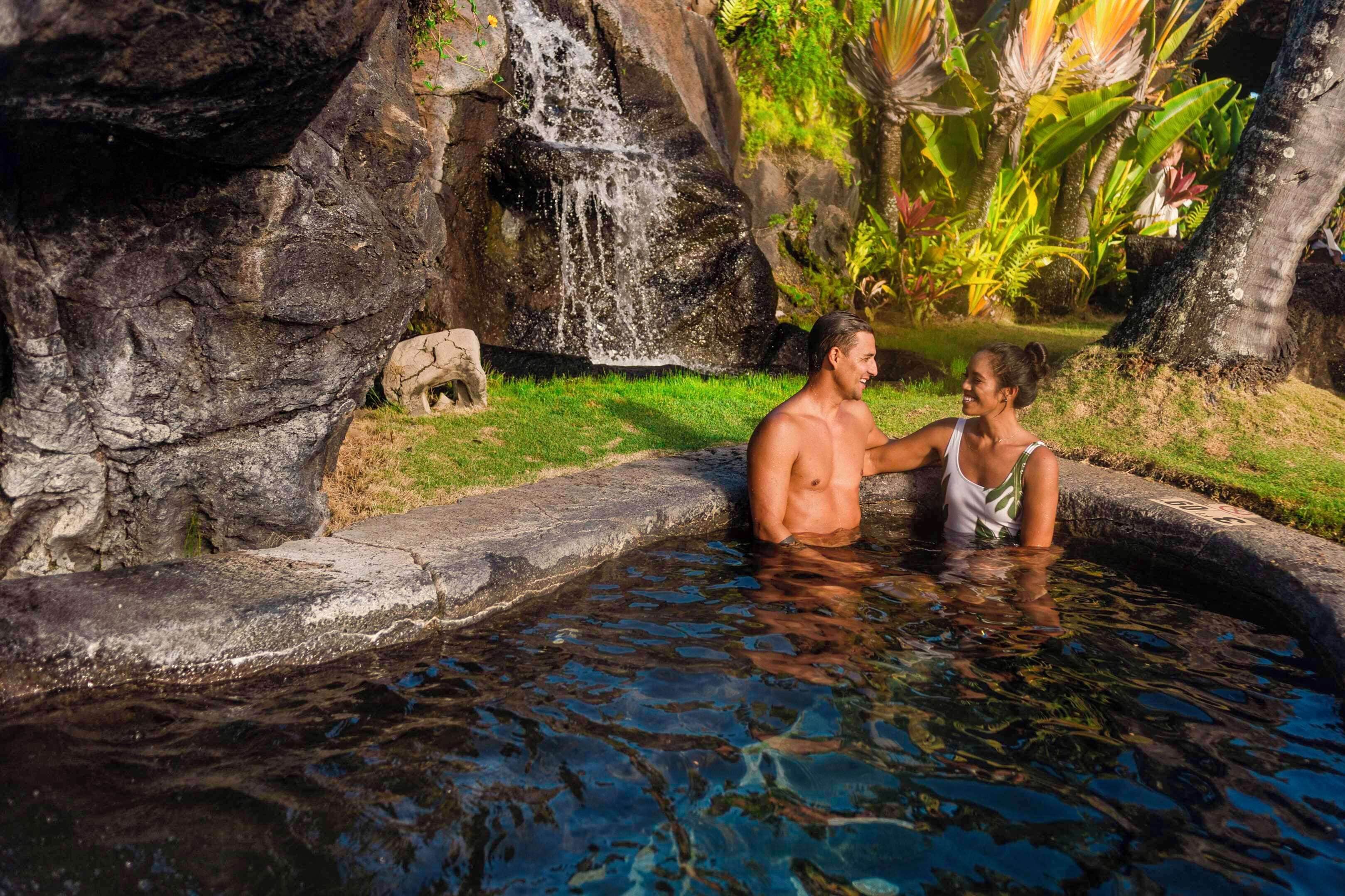 Um homem e uma mulher em uma piscina natural com cascata e plantas tropicais.