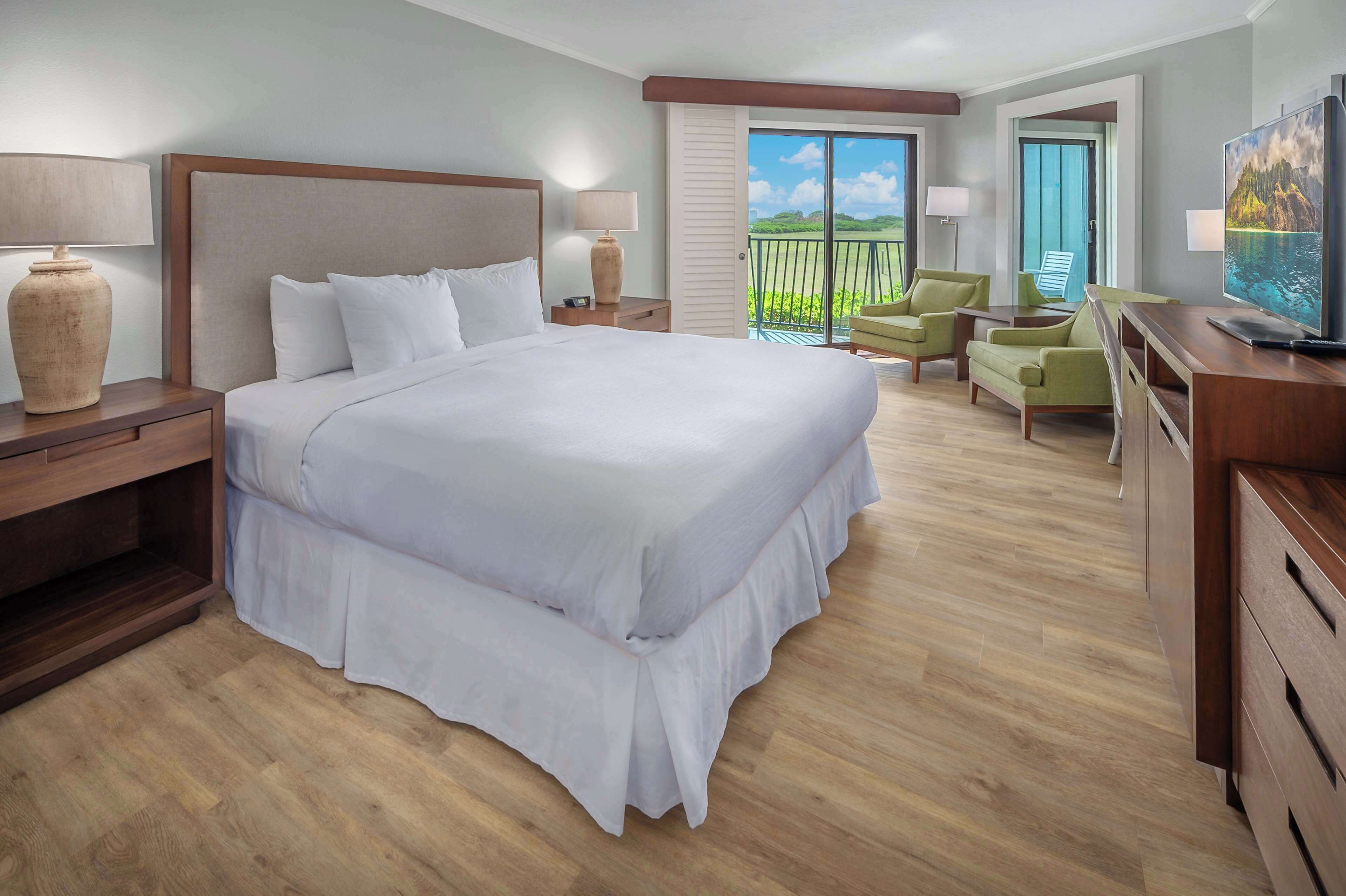 Quarto de hotel com uma cama grande, televisão, varanda com cadeiras e vista para o exterior.