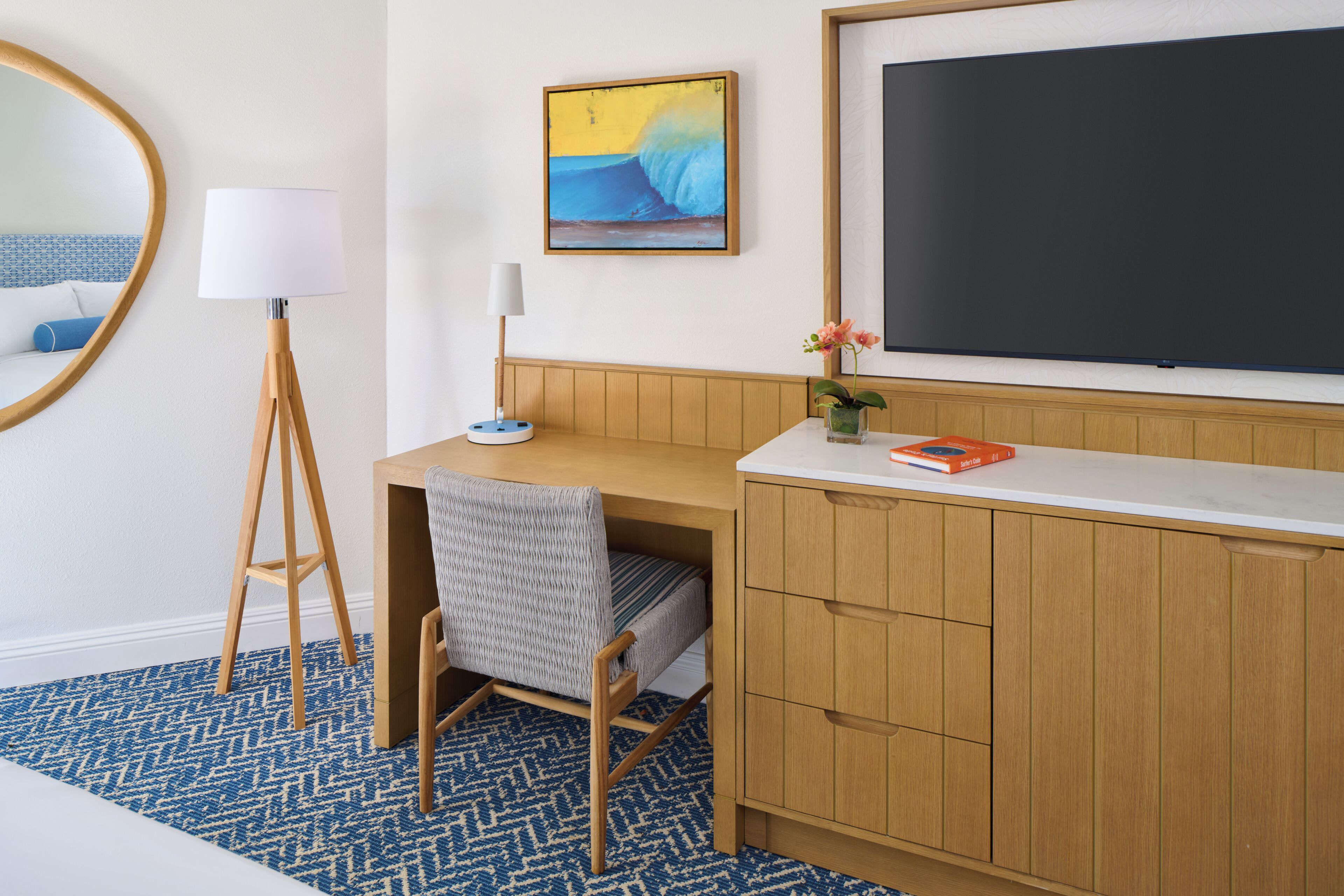 Una camera d'albergo con una scrivania in legno, una sedia, una lampada da terra, un quadro incorniciato, una pianta in vaso e una grande TV a schermo piatto.