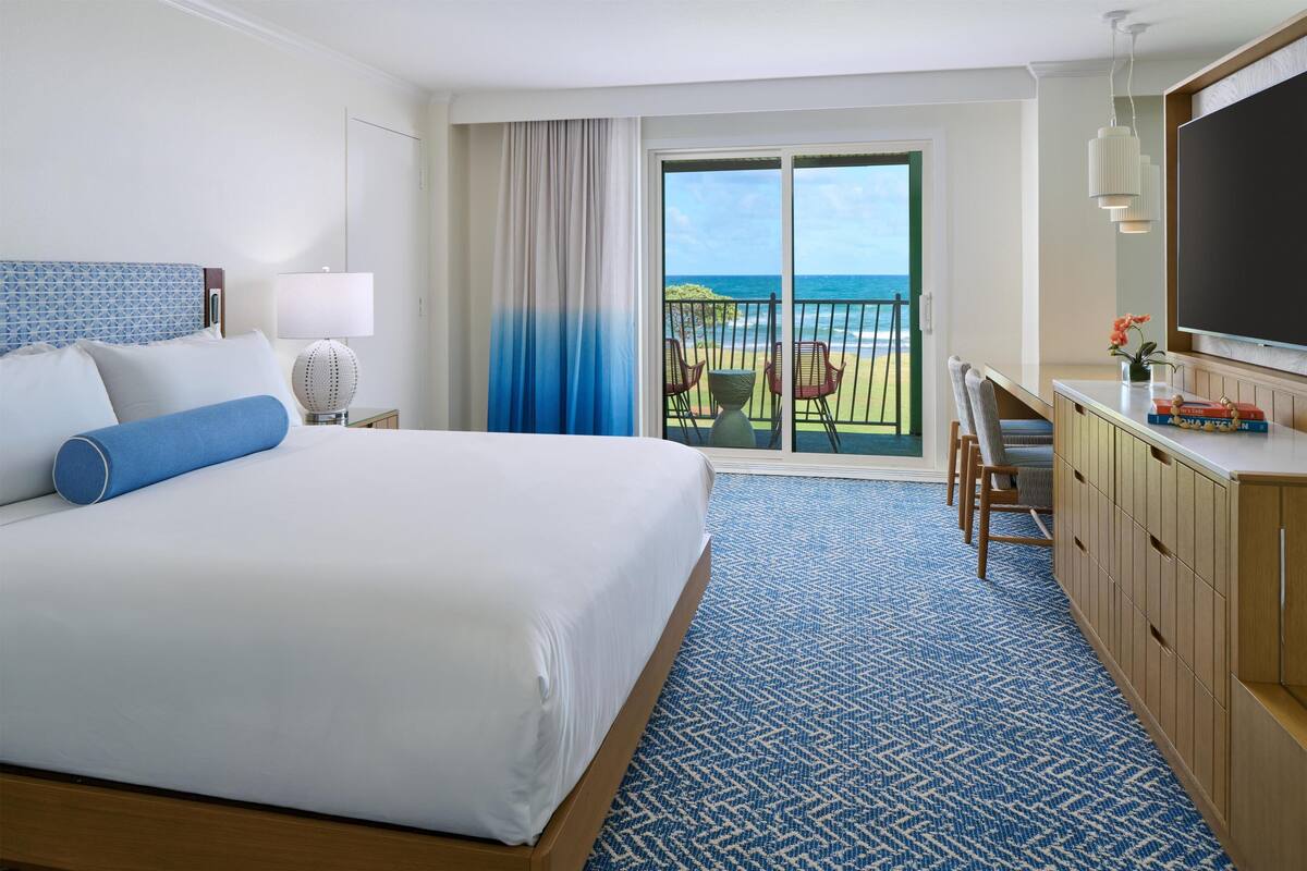 A hotel room with a bed, a desk, a TV, a balcony with a view of the ocean, and a patterned carpet.