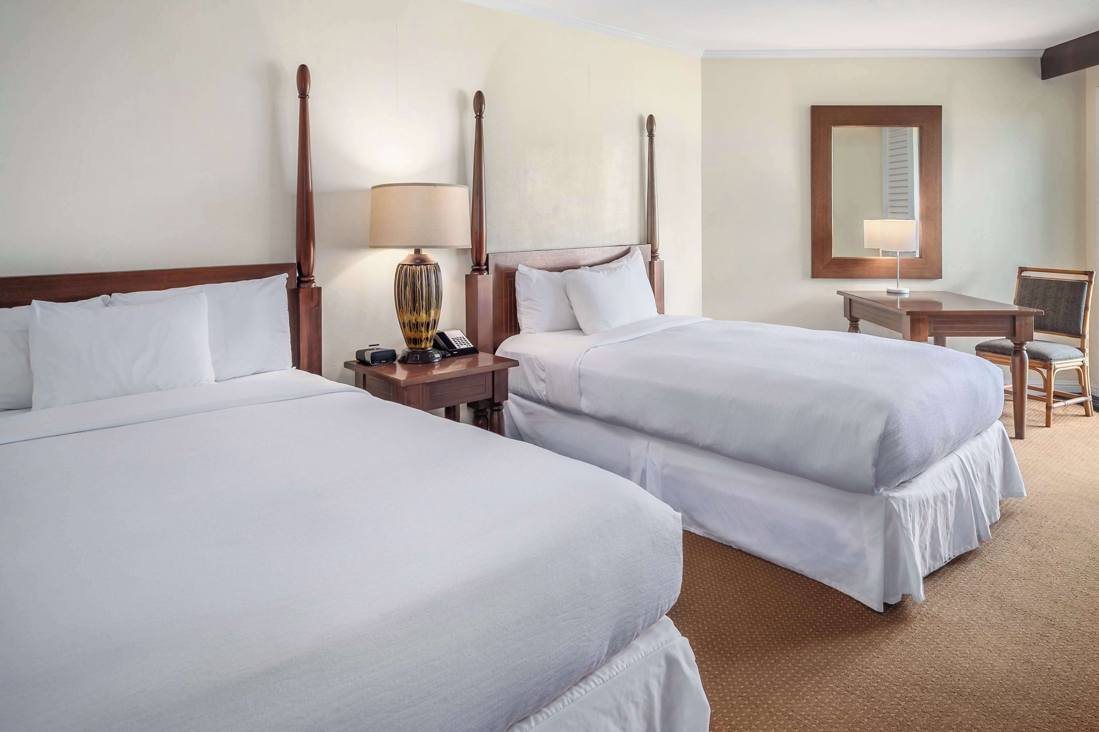Quarto de hotel com duas camas, uma mesa, uma cadeira, um abajur e um espelho.