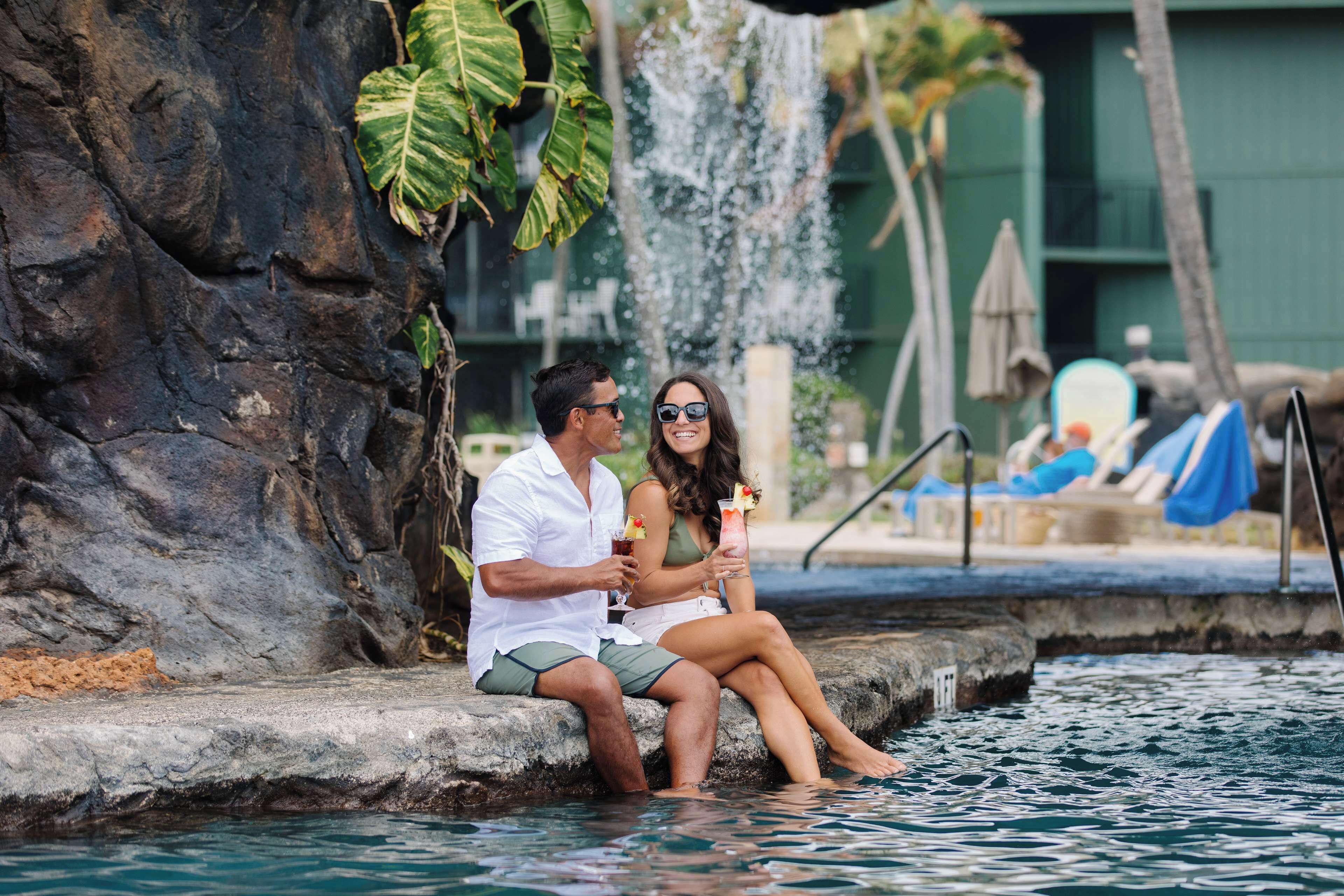 Una pareja sentada junto a una piscina con cascada, rodeada de vegetación y un edificio al fondo.