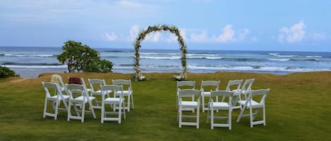A wedding ceremony setup with white chairs arranged in rows facing a decorated arch over a grassy area with a view of the ocean.