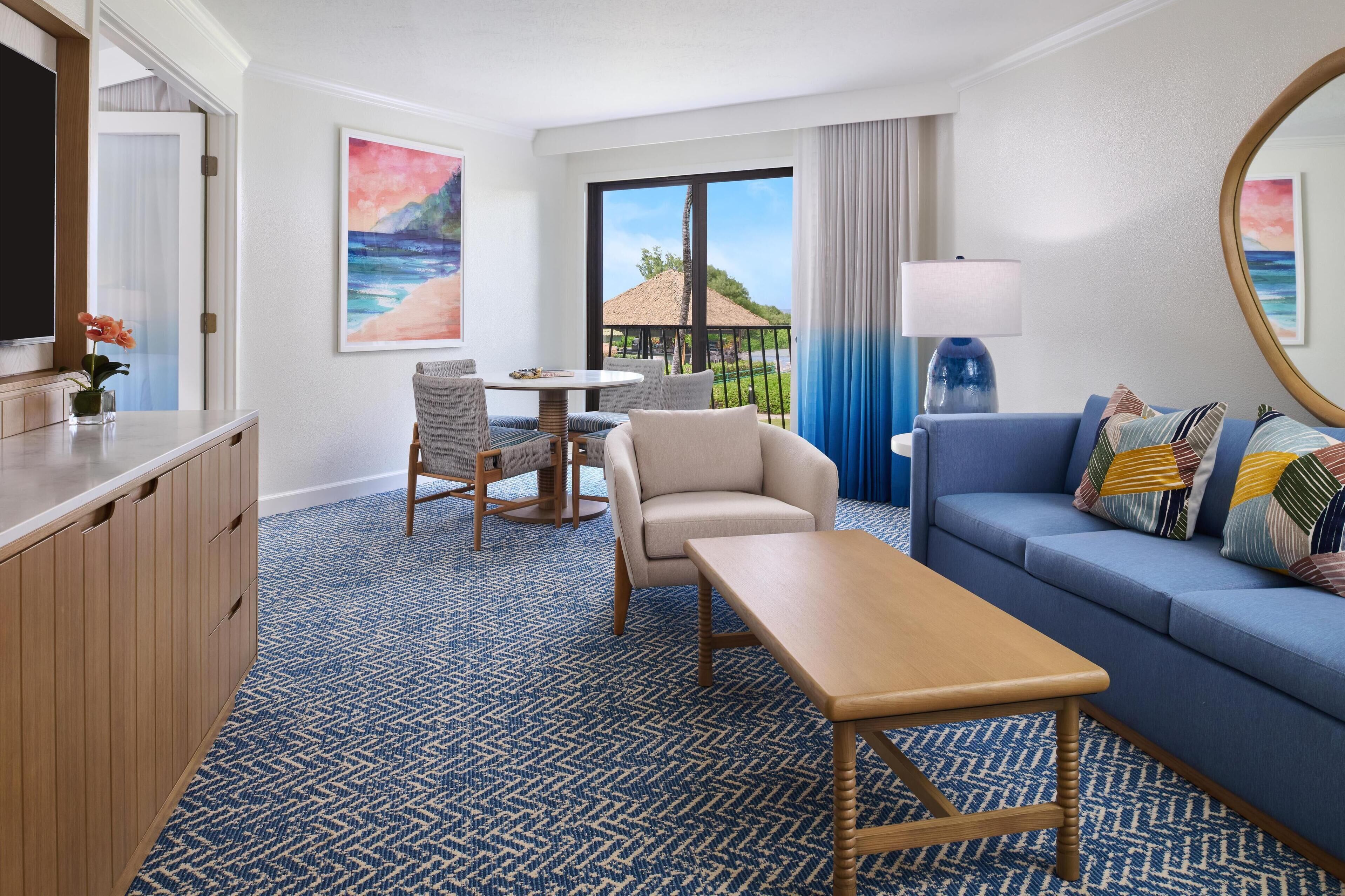 Habitación de hotel con sofá, una mesa de comedor con sillas, televisión y un amplio ventanal con vista a la playa, además de una alfombra azul.