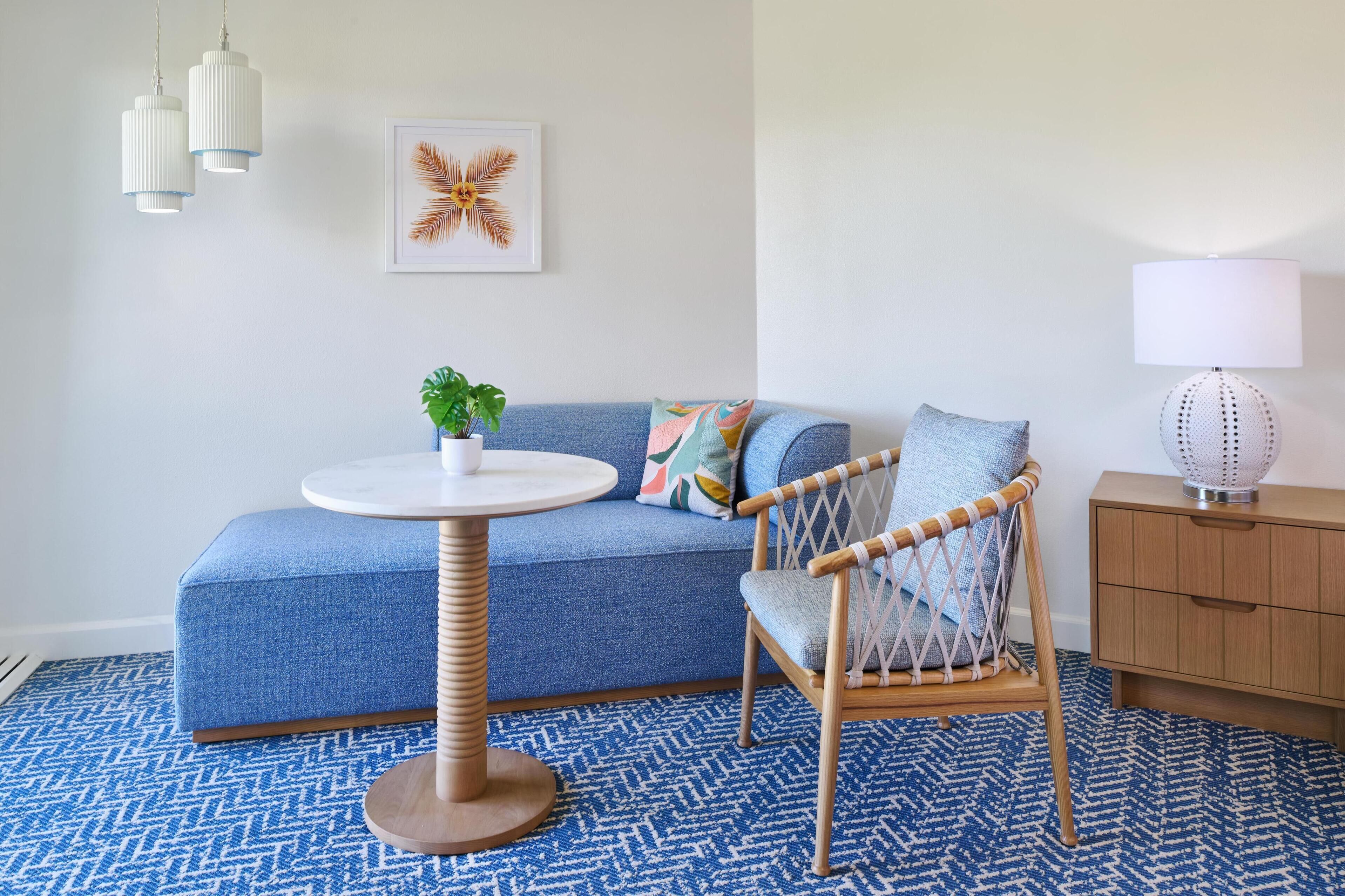 A room with a blue sofa, a wicker chair, a round table with a plant, a lamp, and a framed picture on the wall.
