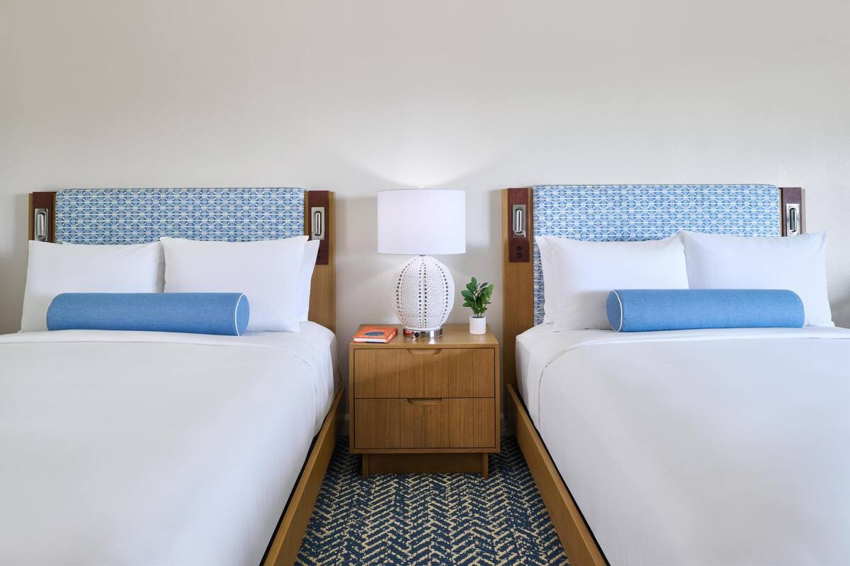 A hotel room with two beds, a nightstand, a lamp, and a small plant.