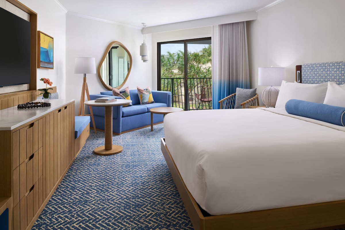 Une chambre d’hôtel avec un lit, un canapé, une petite table, une chaise, une télévision, un miroir et un balcon donnant sur des palmiers.
