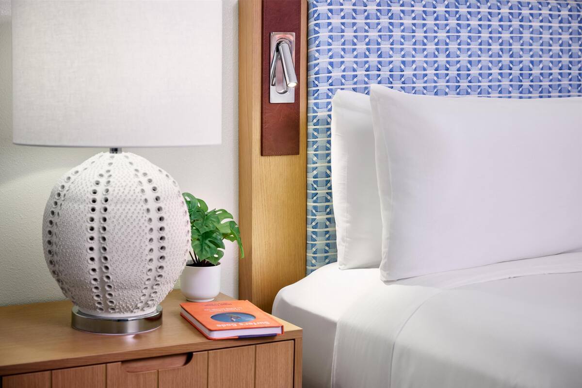 A neatly made bed with white bedding, a bedside lamp, a book, and a potted plant on a wooden nightstand.