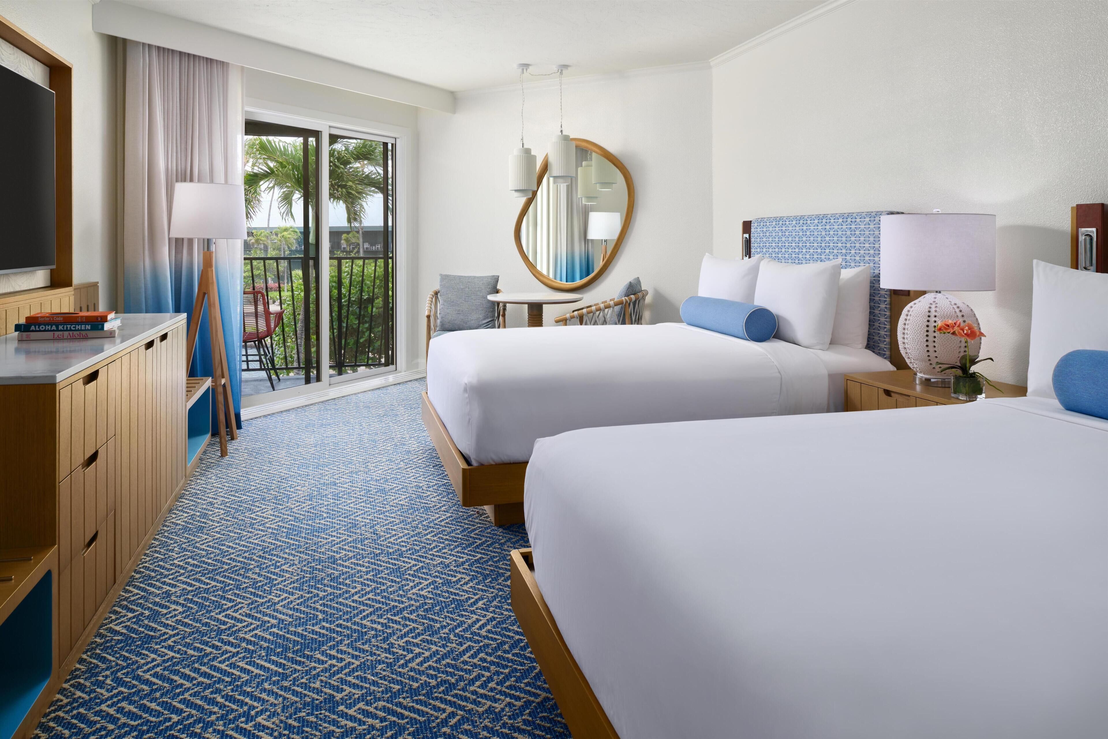 A hotel room with two beds, a TV, a desk, a chair, a mirror, and a view of the outdoors.