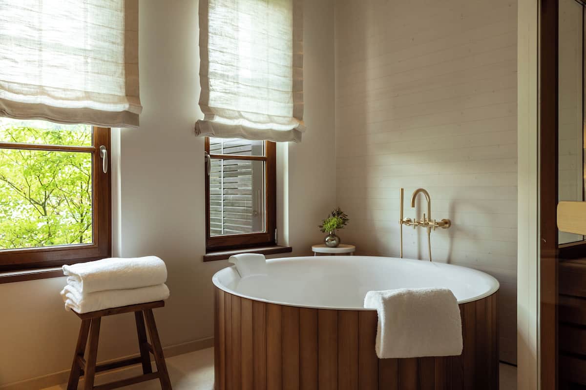 A modern bathroom with a freestanding bathtub, a window with blinds, and a small stool with a towel.