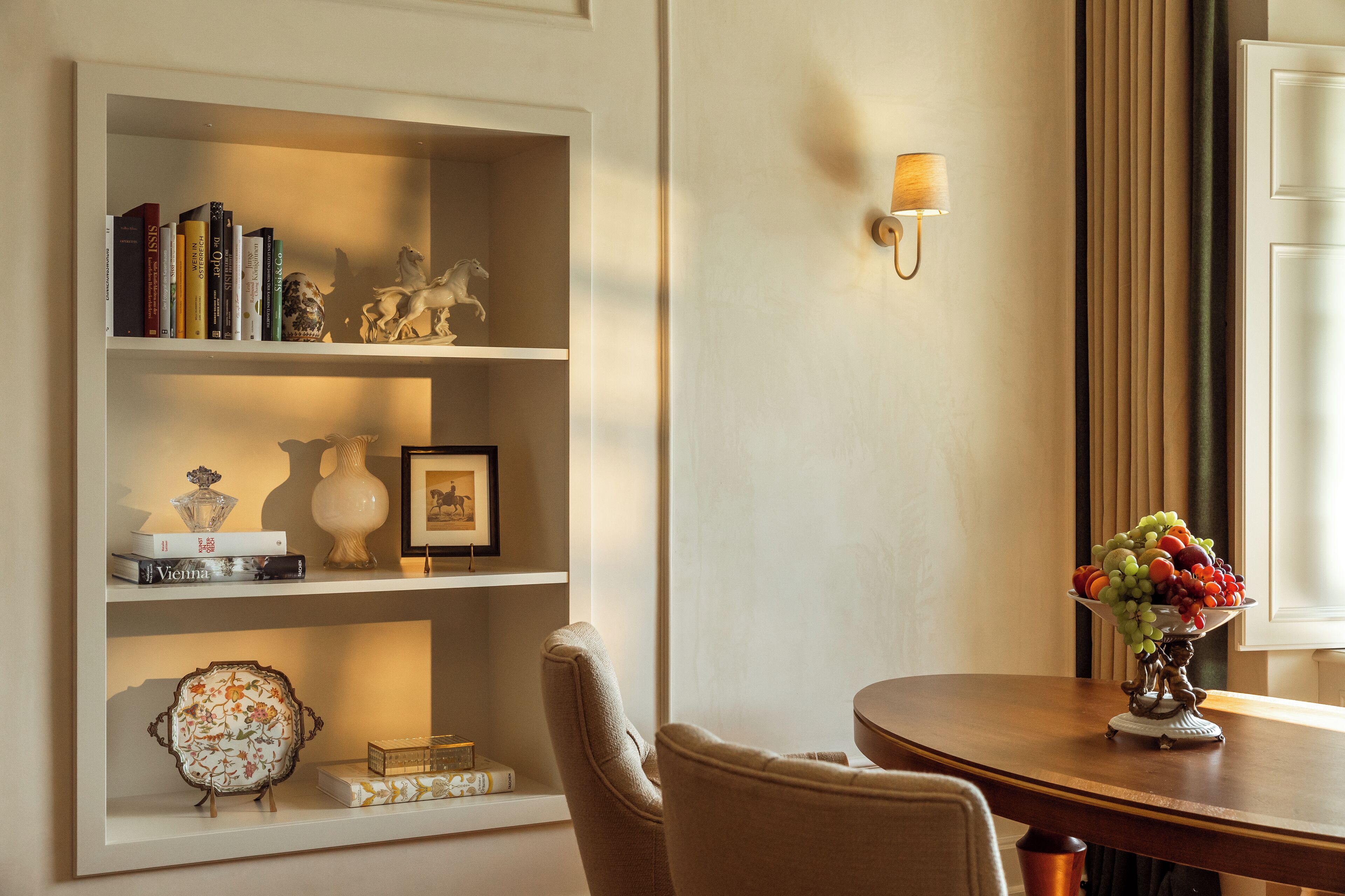 Uma sala de jantar com mesa de madeira, uma cadeira, estante com livros e objetos decorativos, e luminária fixada na parede.