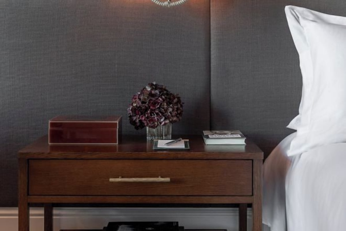 Una mesita de noche de madera con un cajón, un florero con flores y un libro.