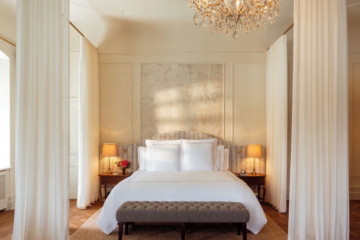 A spacious bedroom with a large bed, two bedside tables with lamps, a bench, and a chandelier.
