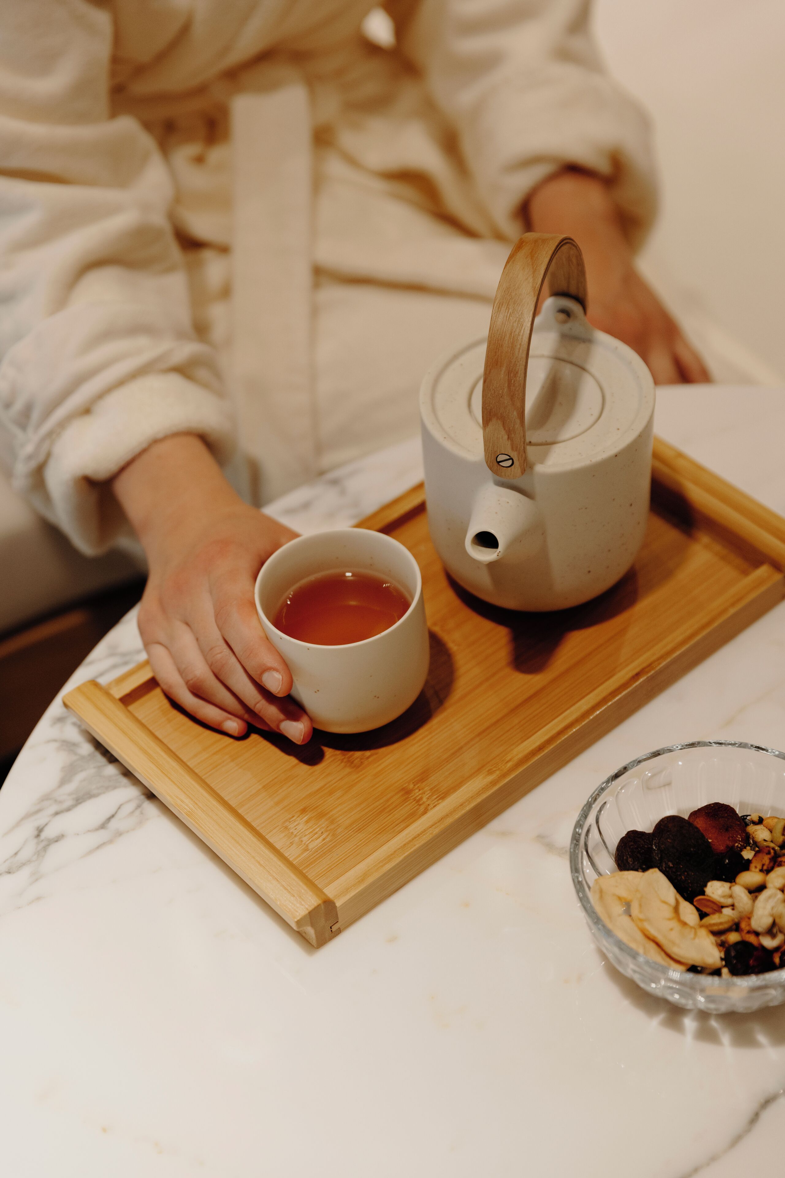 A wooden tray with a teapot and a cup of tea, a bowl of nuts and dried fruits, and a marble countertop.