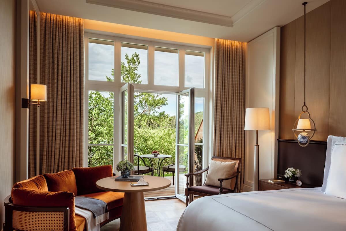 A hotel room with a large bed, a sofa, a round table, and a chair. There are curtains on the windows, and a lamp on the wall.