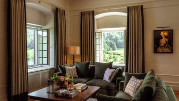 A cozy living room with a green sofa set, a wooden coffee table with a book and flowers, and a view of a lush garden through large windows.