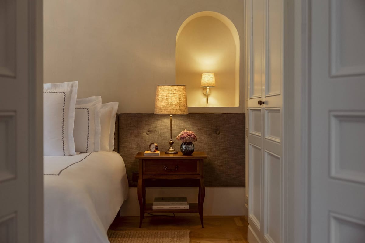 A neatly made bed with white linens and pillows, a bedside table with a lamp, a small clock, and a vase with flowers.