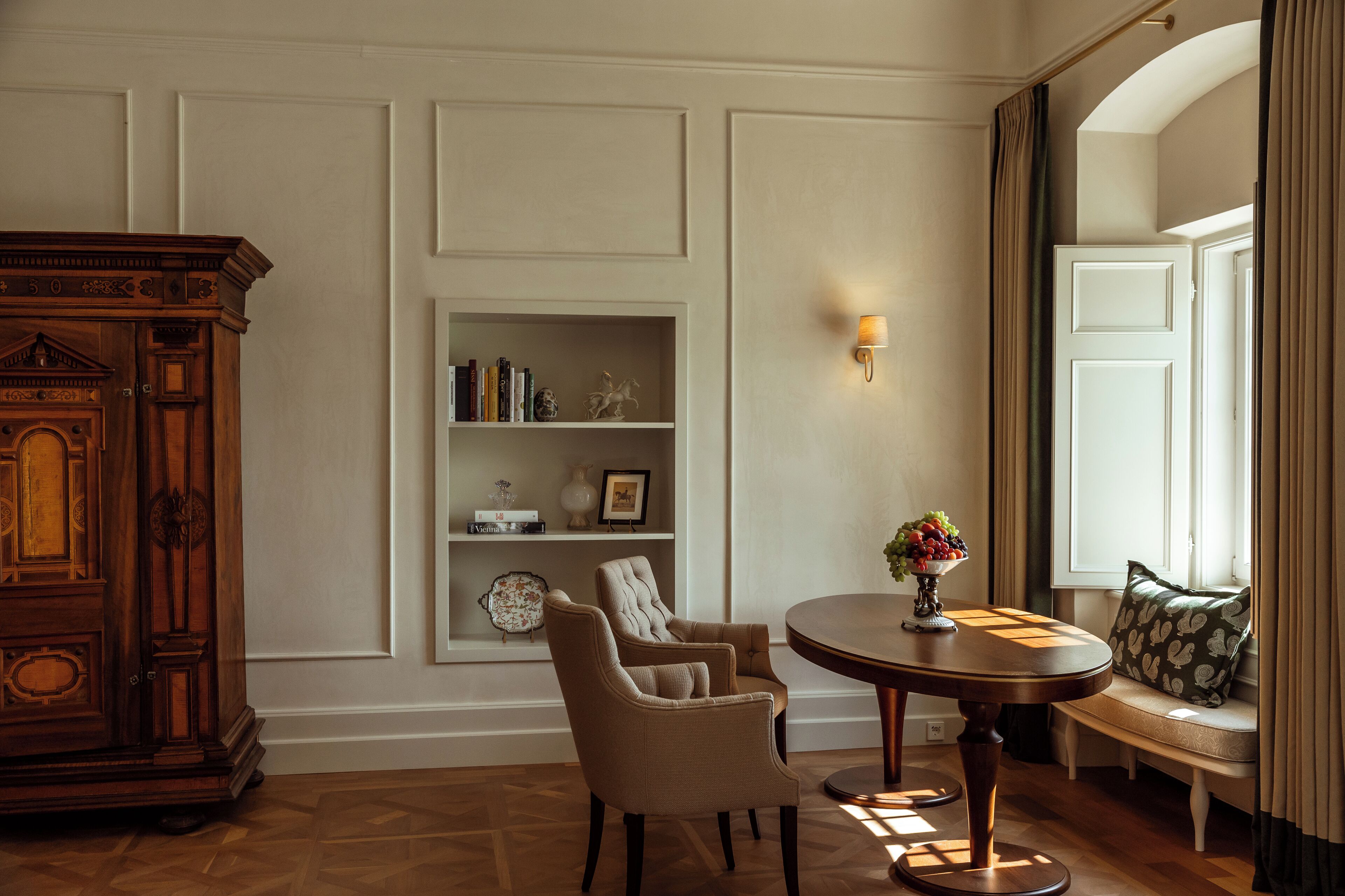A room with a wooden cabinet, two armchairs, a round table with a vase of flowers, a bookshelf, and a window with curtains.