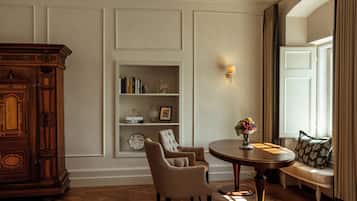 A room with a wooden cabinet, two armchairs, a round table with a vase of flowers, a bookshelf, and a window with curtains.