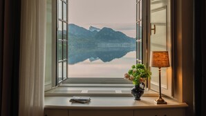 Janela com vista para montanhas e um lago, uma luminária e um vaso com flores sobre uma escrivaninha.