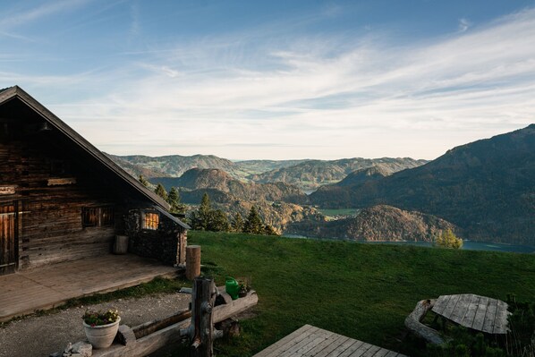 Una cabaña rústica con un porche de madera, rodeada de un paisaje verde exuberante y montañas al fondo.