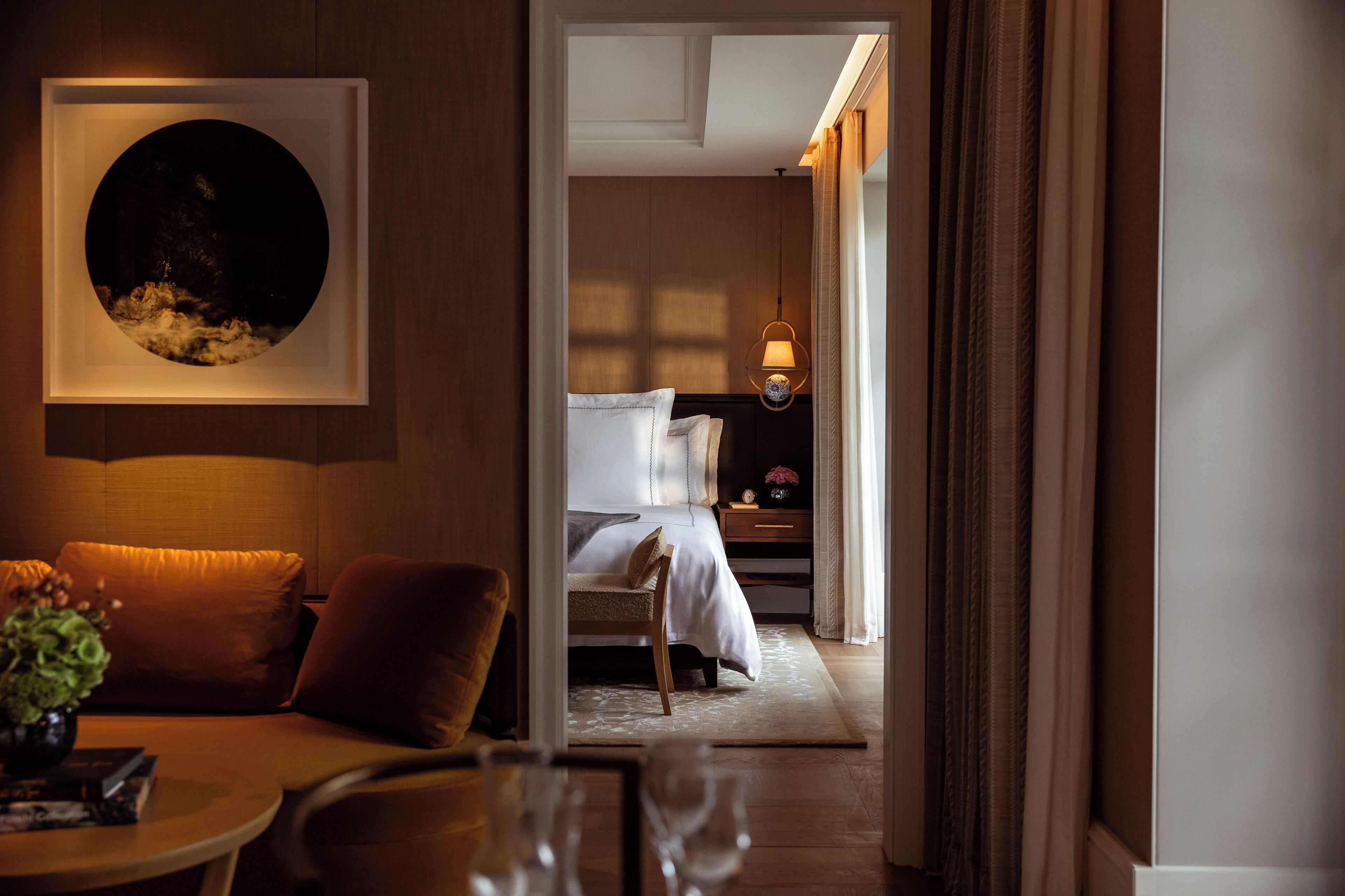 Habitación de hotel con cama, mesita de noche, lámpara y vista a otra habitación.
