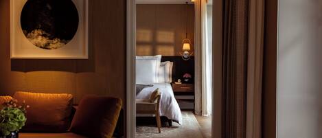 Habitación de hotel con cama, mesita de noche, lámpara y vista a otra habitación.
