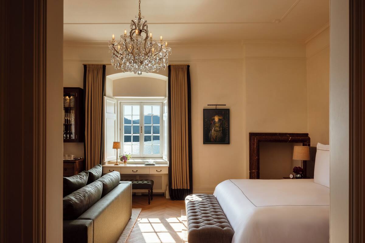 A hotel room with a large bed, a sofa, a desk with a lamp, a chandelier, and a window with curtains.