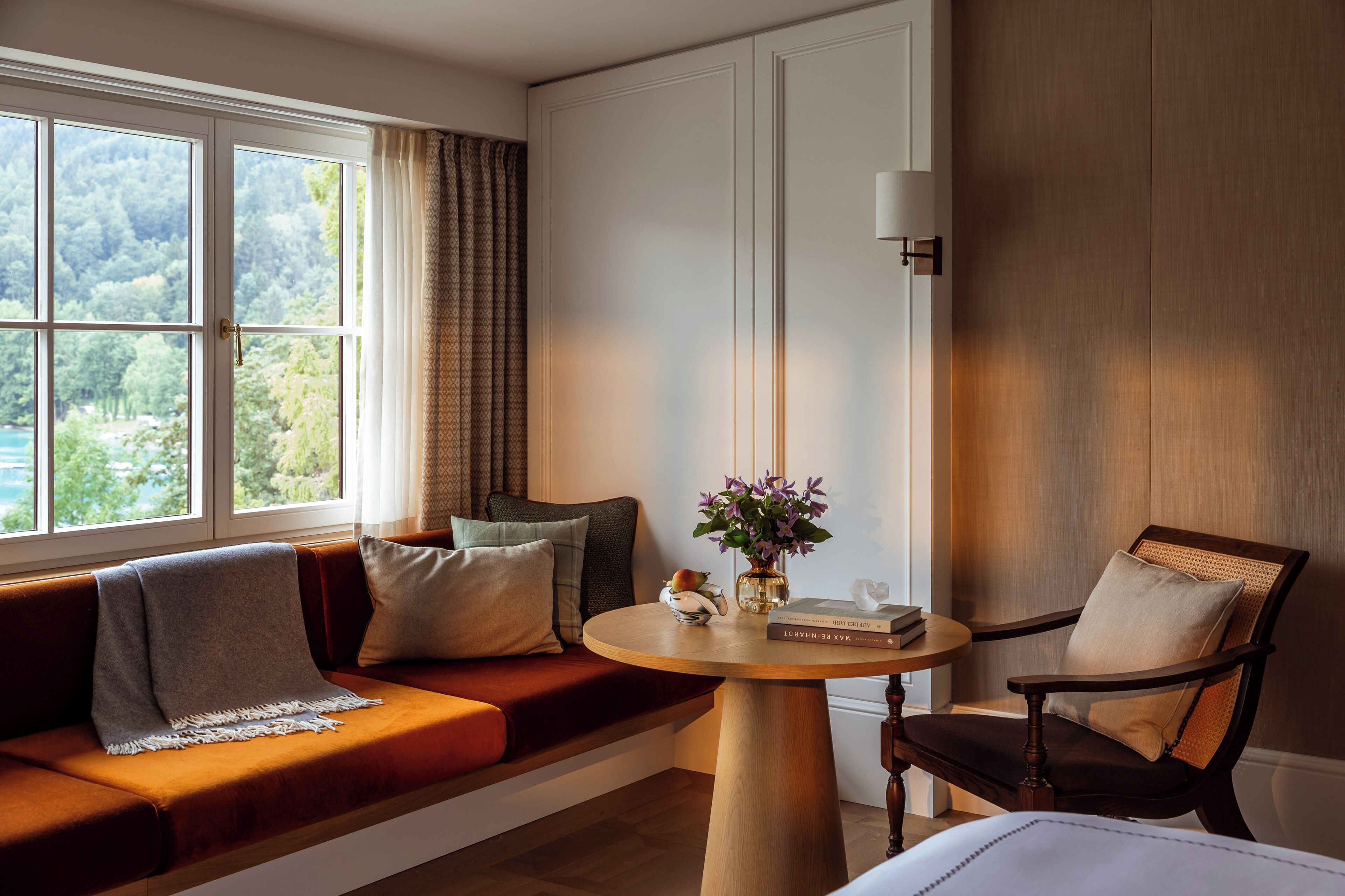 A cozy room with a wooden bench, a round table with a book and flowers, and a view of greenery through a window.
