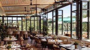Uma ampla área de refeições interna com várias mesas e cadeiras, grandes janelas com vista para o exterior, e um bar com prateleiras cheias de garrafas.
