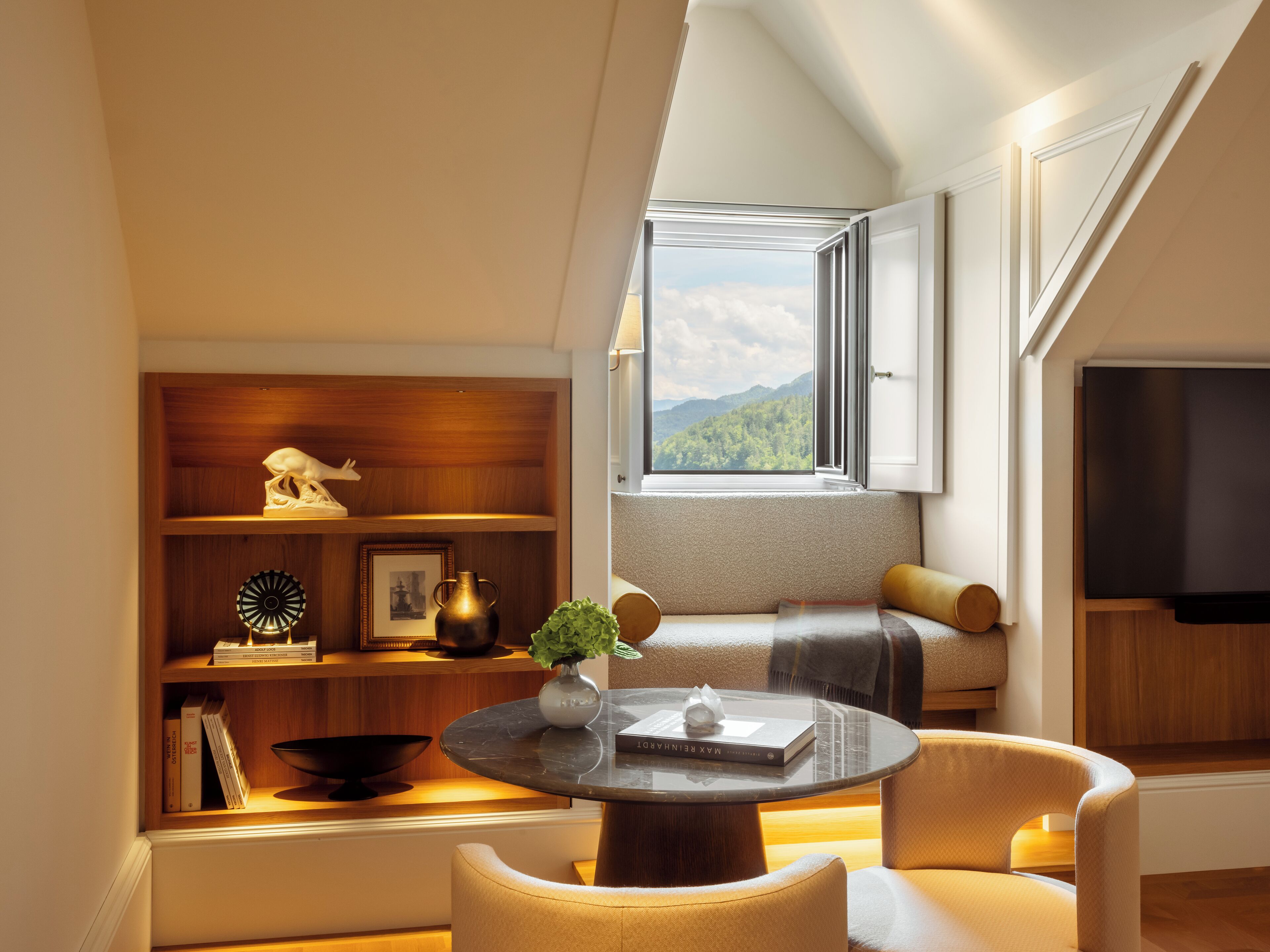 Een moderne woonkamer met een ronde tafel, zachte zitplaatsen, een boekenkast en een groot raam met uitzicht op bergen.