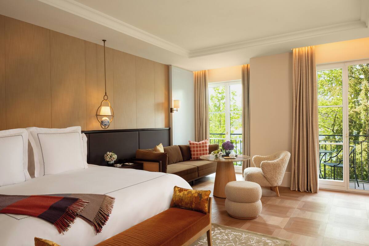 A hotel room with a large bed, a sofa, a small table, and a balcony with a view of trees.