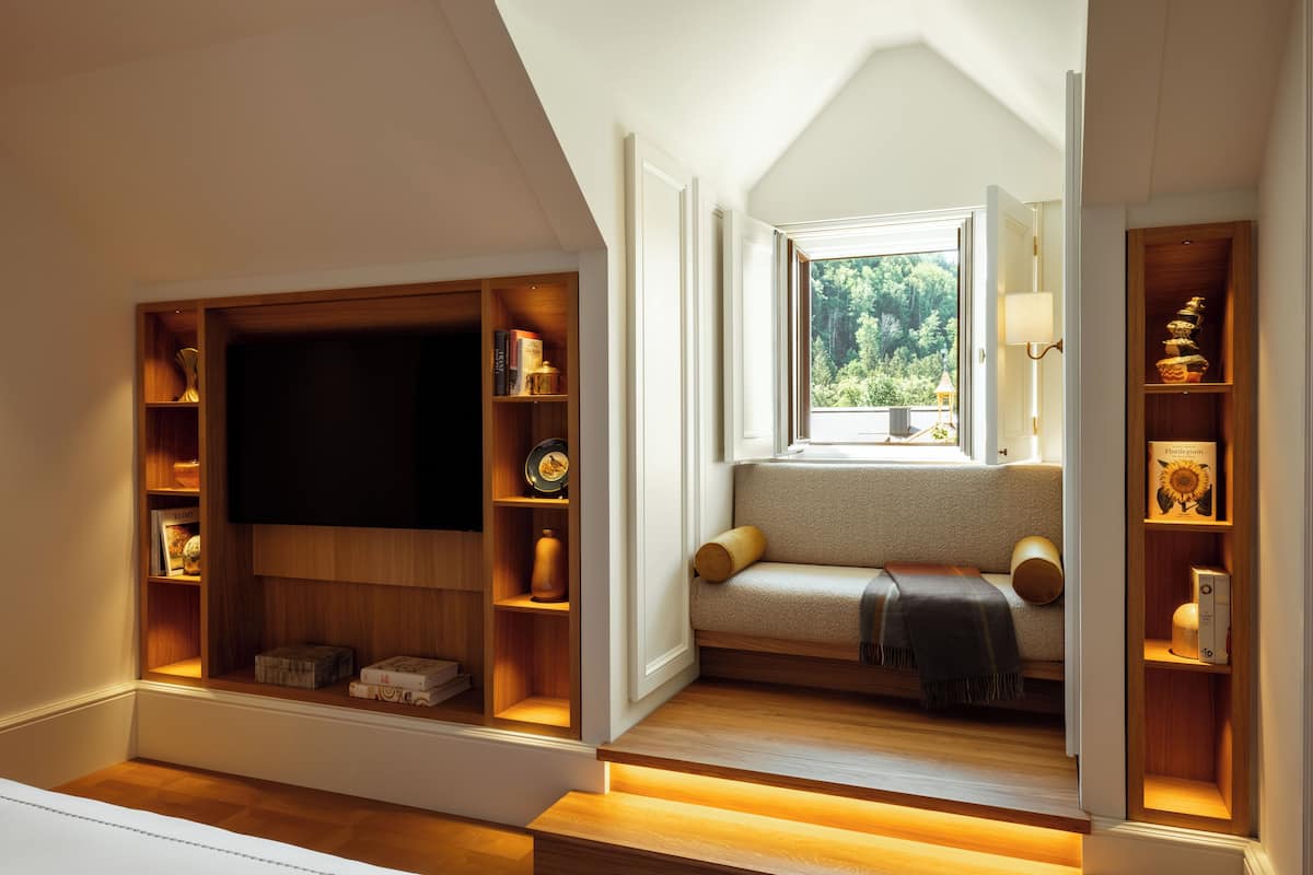 A cozy room with a built-in TV, wooden shelves, a comfortable seating area with a view of greenery outside.