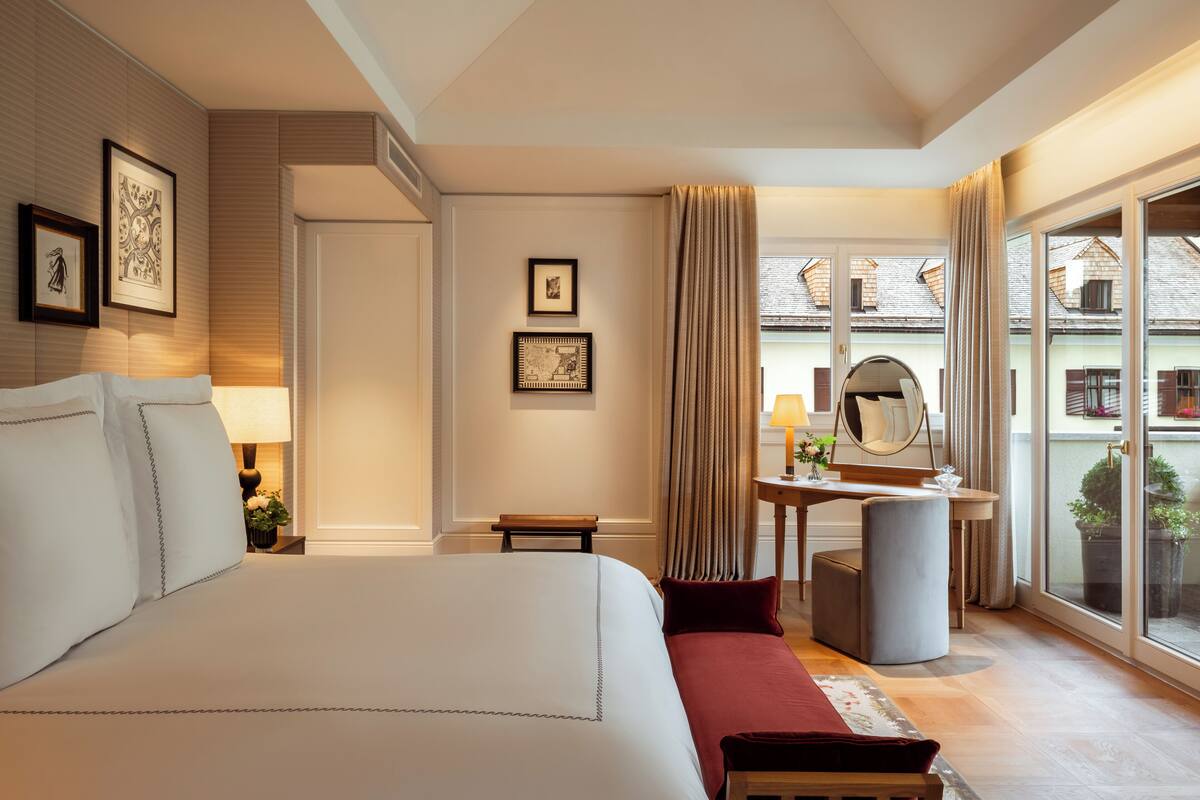 A hotel room with a large bed, a desk with a mirror, a chair, and a view of buildings through large windows.