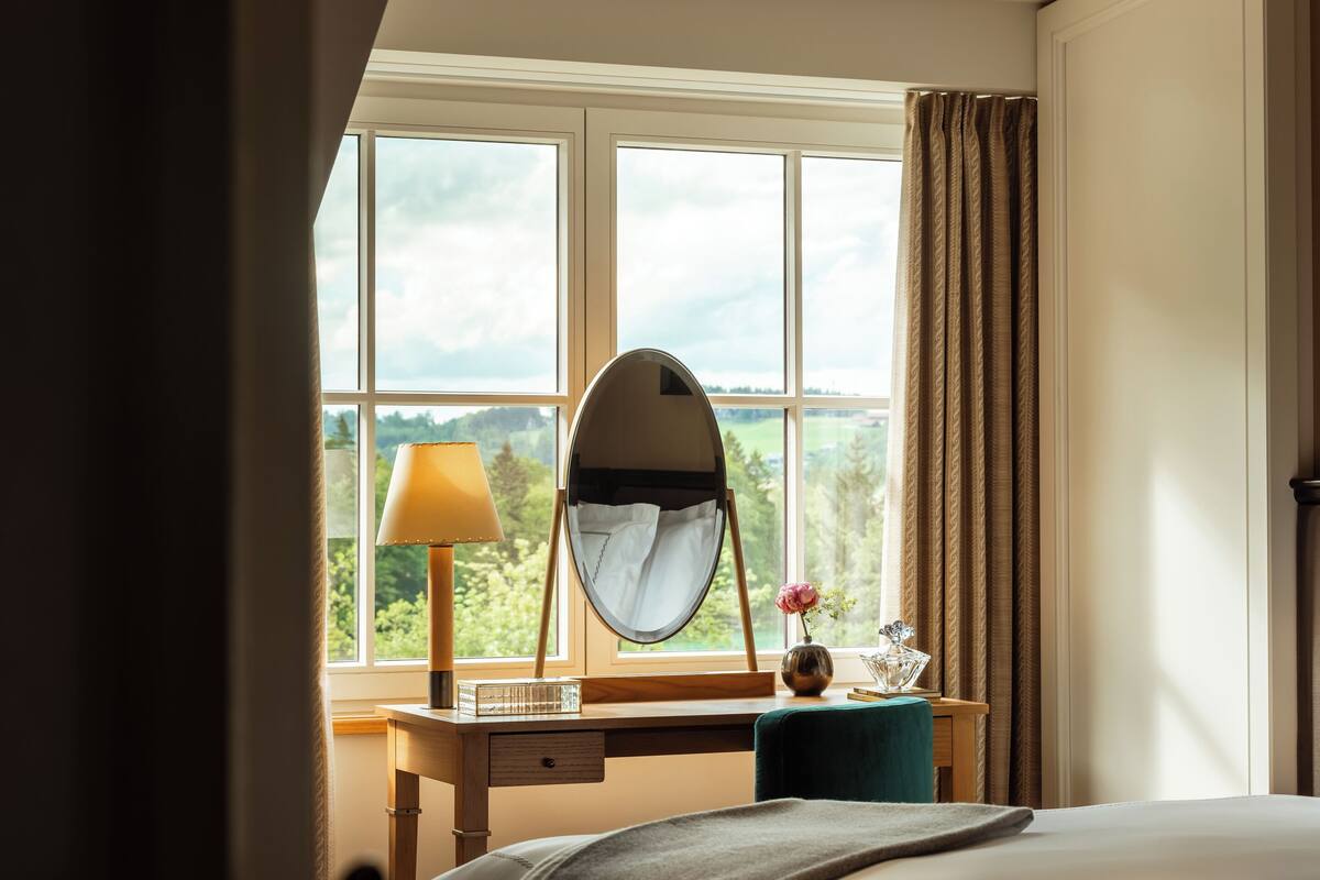 A bedroom with a large window, a bedside table with a lamp, a mirror, and a vase with flowers.