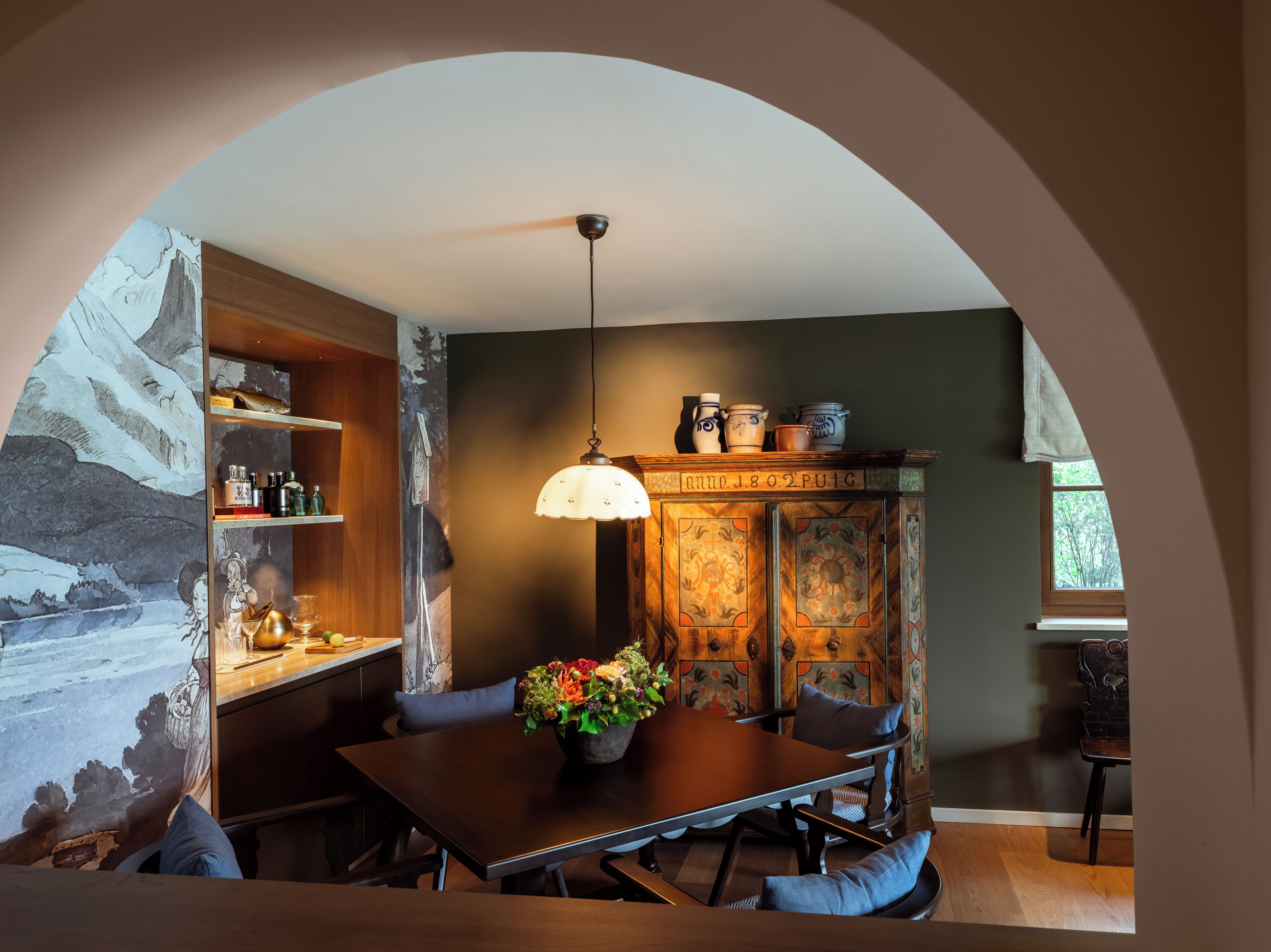 A dining area with a table, chairs, a hanging light fixture, and a decorative cabinet with a window view.