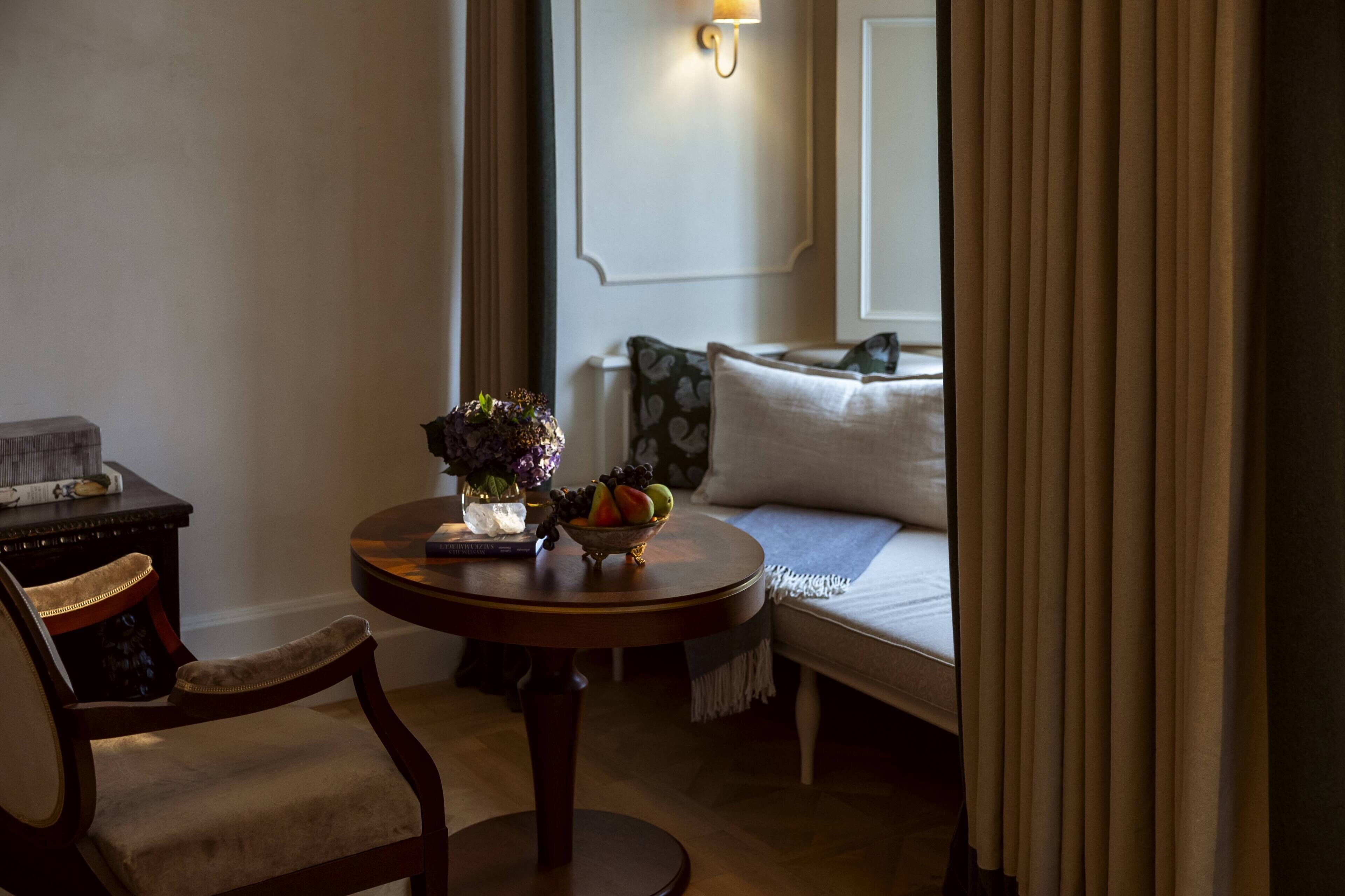 A cozy room with a round wooden table, a chair, a sofa with patterned cushions, and a vase with flowers.