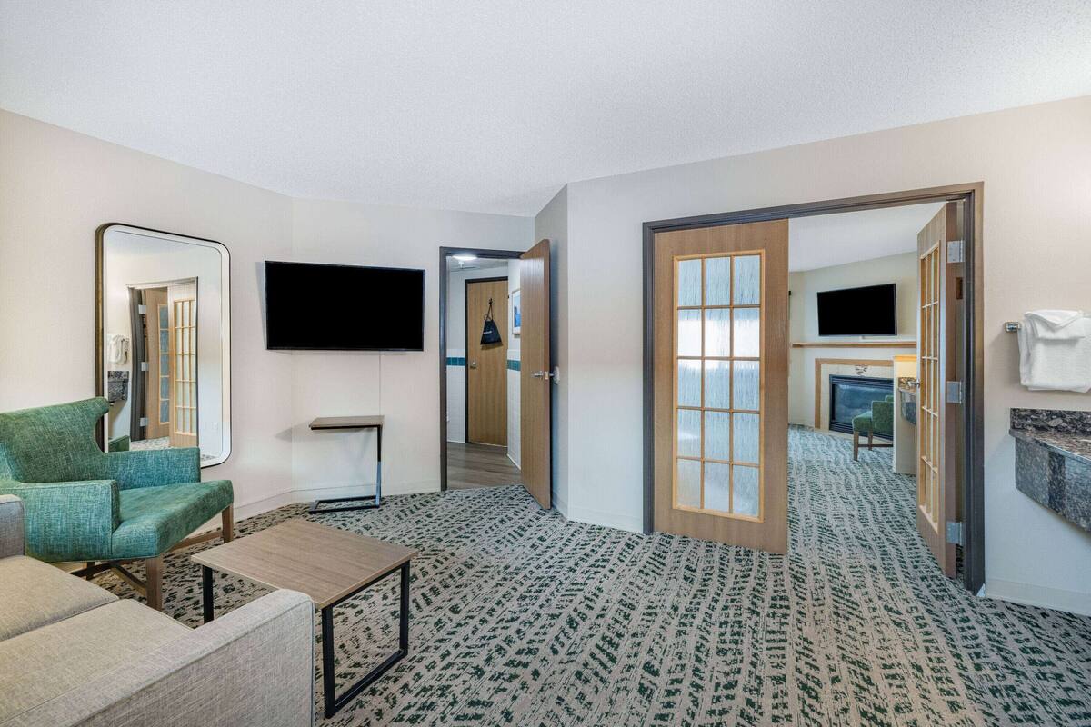 Een hotelkamer met een groene fauteuil, een klein tafeltje, een flatscreen televisie, een open haard en een spiegel.